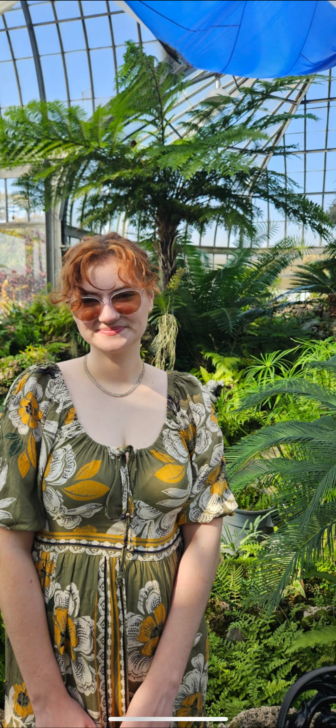 A young woman with short, curly red hair wearing sunglasses and a green floral dress standing in a lush greenhouse filled with various green plants and ferns.
