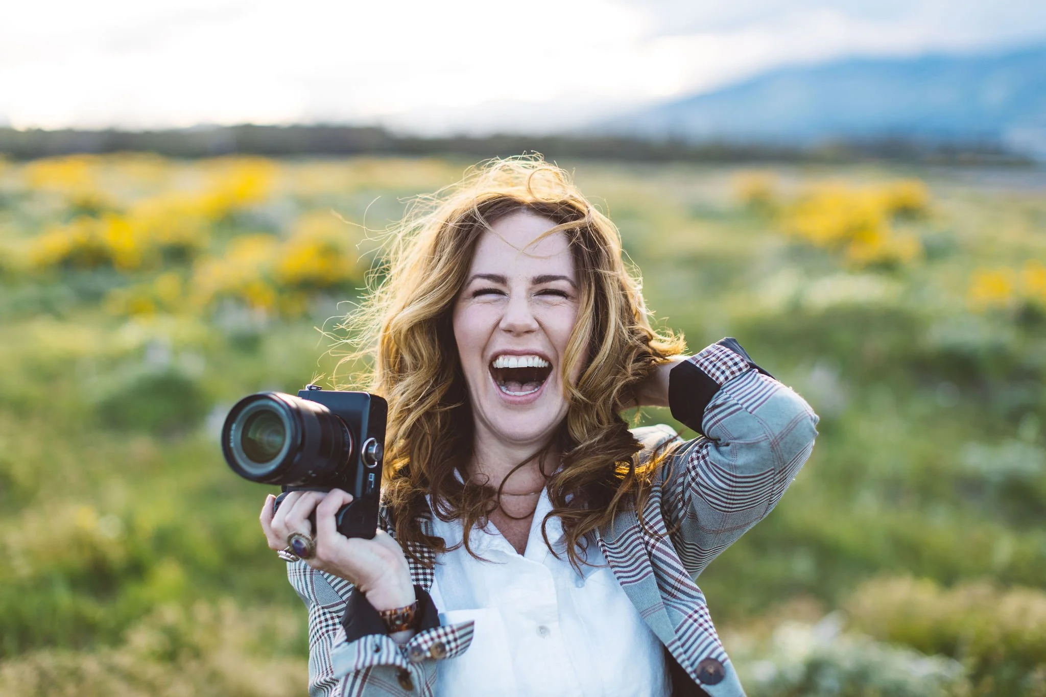 Creative portrait of a woman professional photographer business owner with reddish hair laughing and holding a camera outdoors with a blurred green field and mountains in the background, during a branding photoshoot in Boise, Idaho.