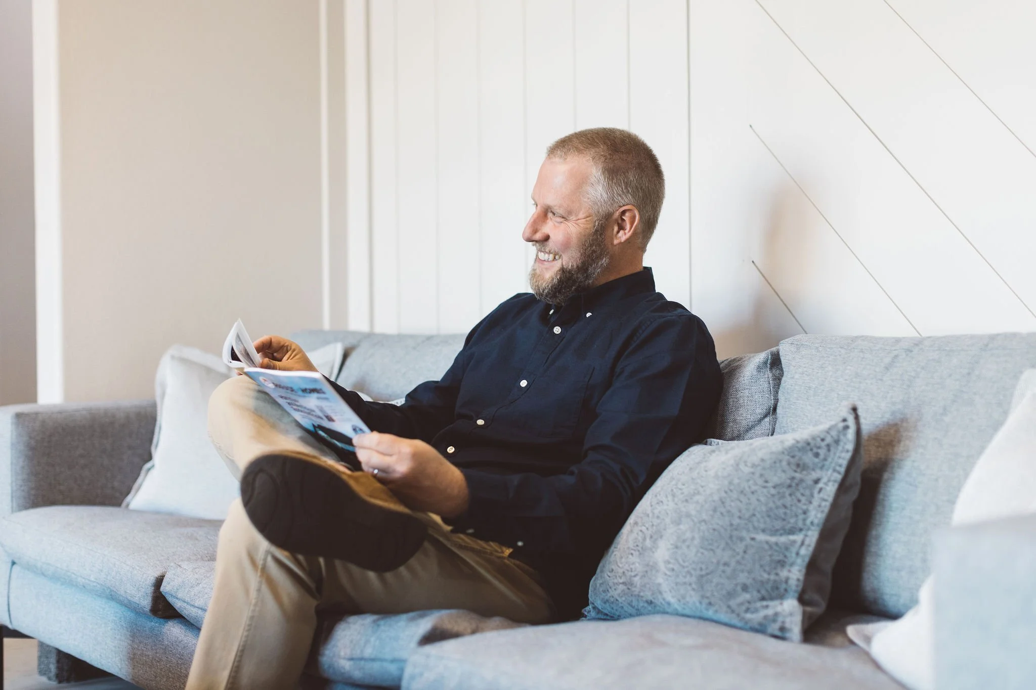 A man with a beard and short hair sitting on a light gray sofa, smiling and reading a magazine in a room with white walls.