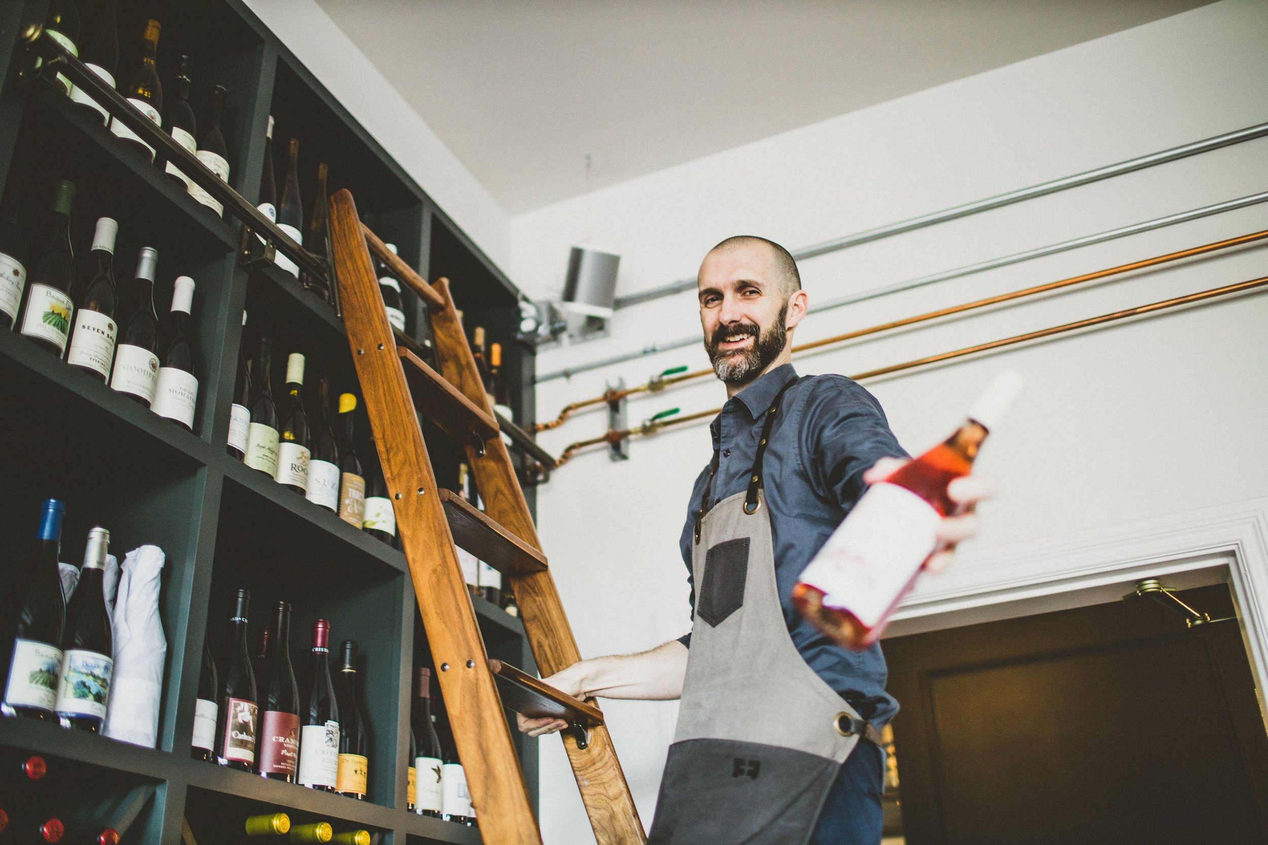 A man in a grey apron holding a bottle of rosé wine inside a wine shop.