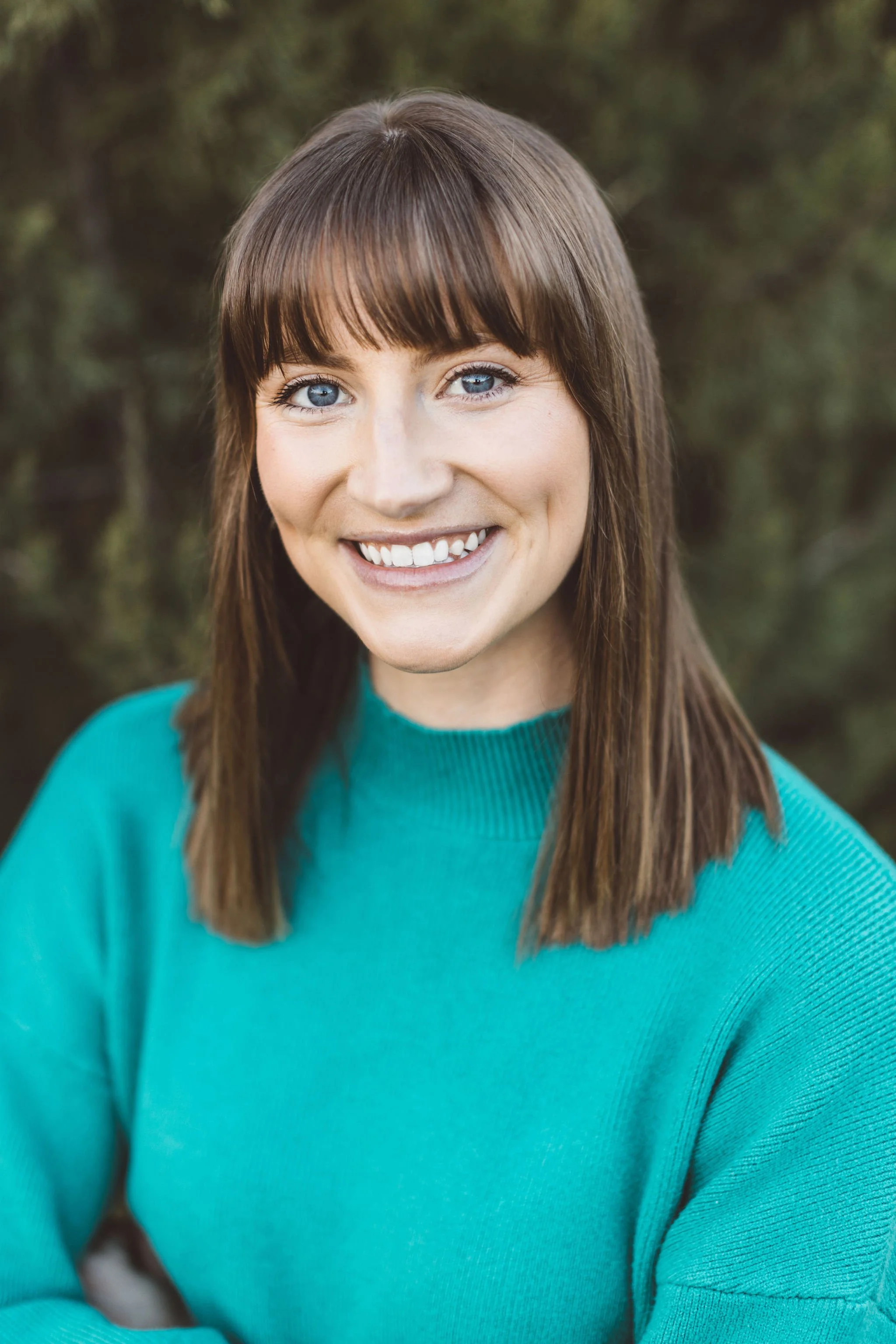 Authentic headshot portrait of a woman with brown hair, blue eyes, smiling, wearing a teal sweater, outdoors in the foothills with blurred greenery in the background during a professional headshot session in Boise, Idaho.
