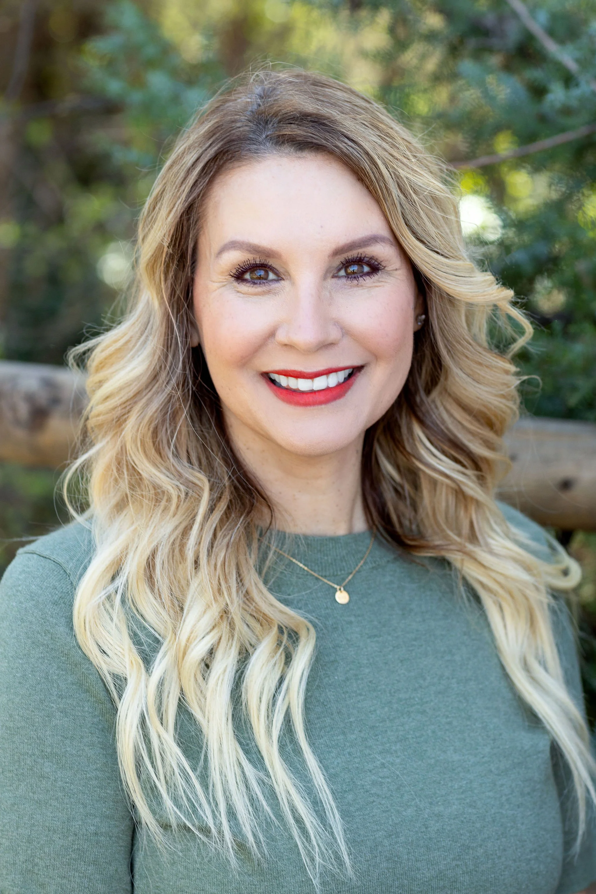 Professional headshot of a woman with wavy blonde hair, wearing a green top and a gold necklace, smiling outdoors in a downtown park with trees in the background during a professional headshot photography session in Boise, Idaho..