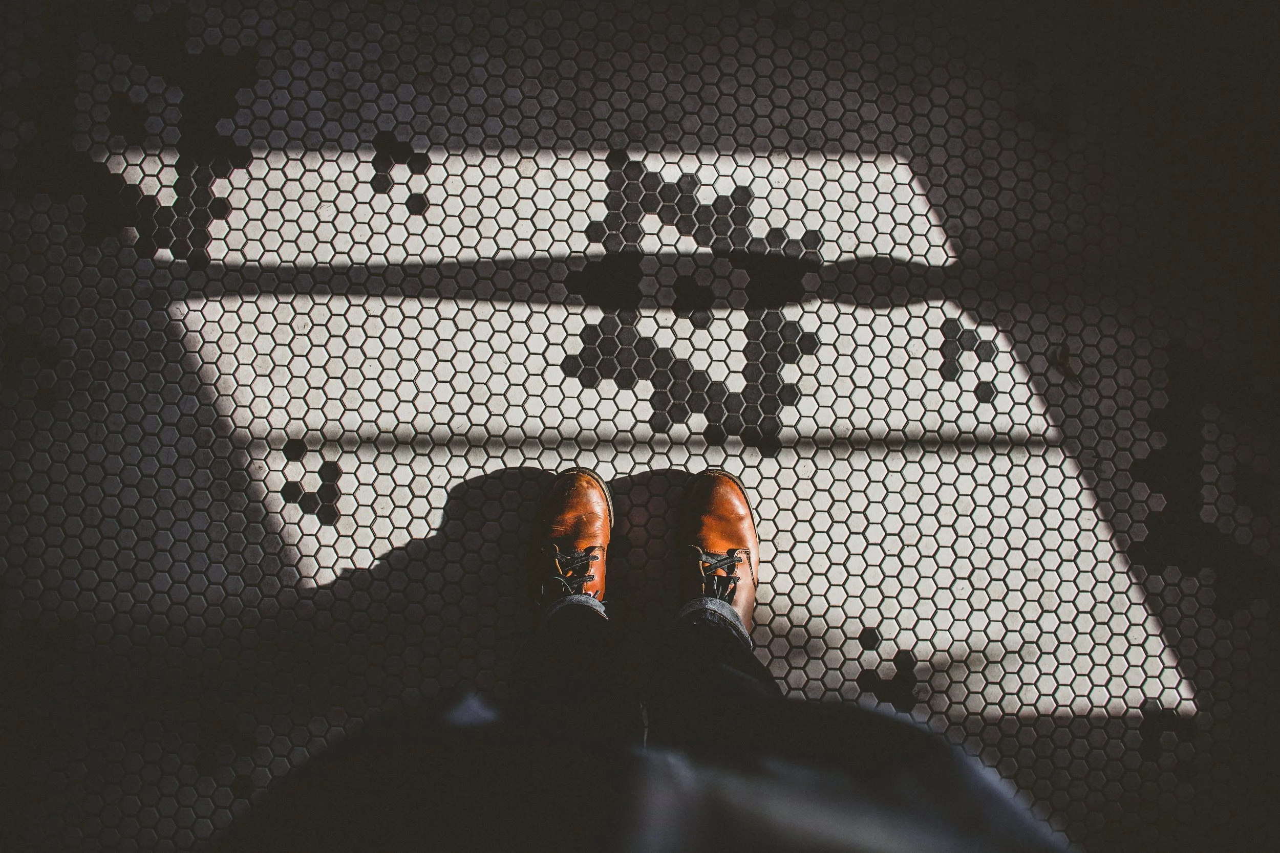 A person's brown shoes and jeans as they stand on a hexagonal-tiled floor, with a large illuminated letter 'A' visible through the tiles.