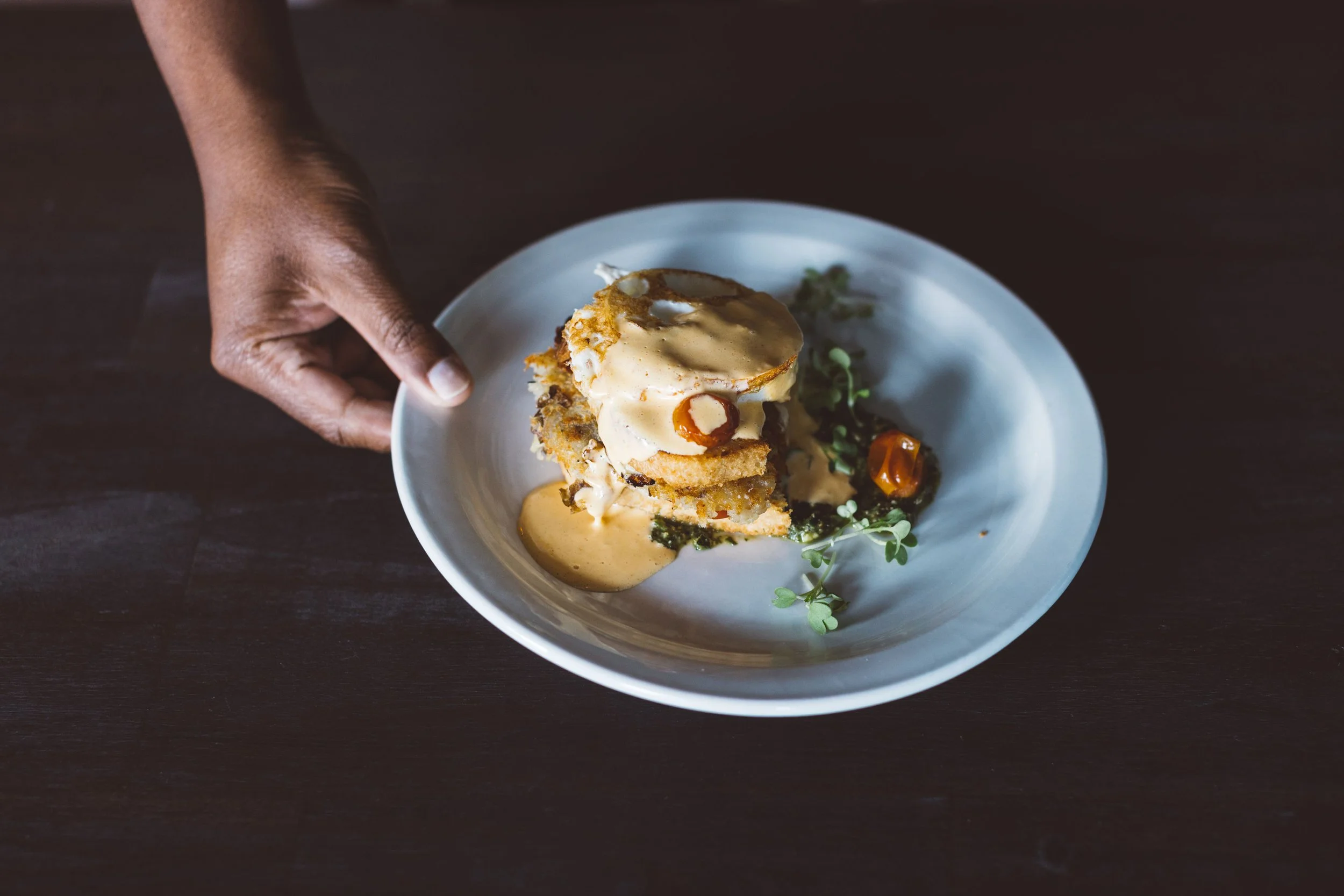 A person's hand holding a white plate with a layered dish topped with sauce, garnished with microgreens, cherry tomatoes, and greens, on a dark wooden table during a branding photo session in Boise, Idaho.