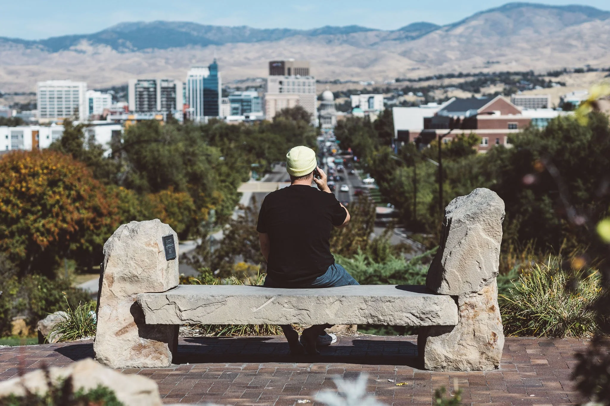 A person sitting on a stone bench, wearing a yellow beanie and black shirt, talking on a cellphone, overlooking a cityscape with buildings, trees, and mountains in the background.