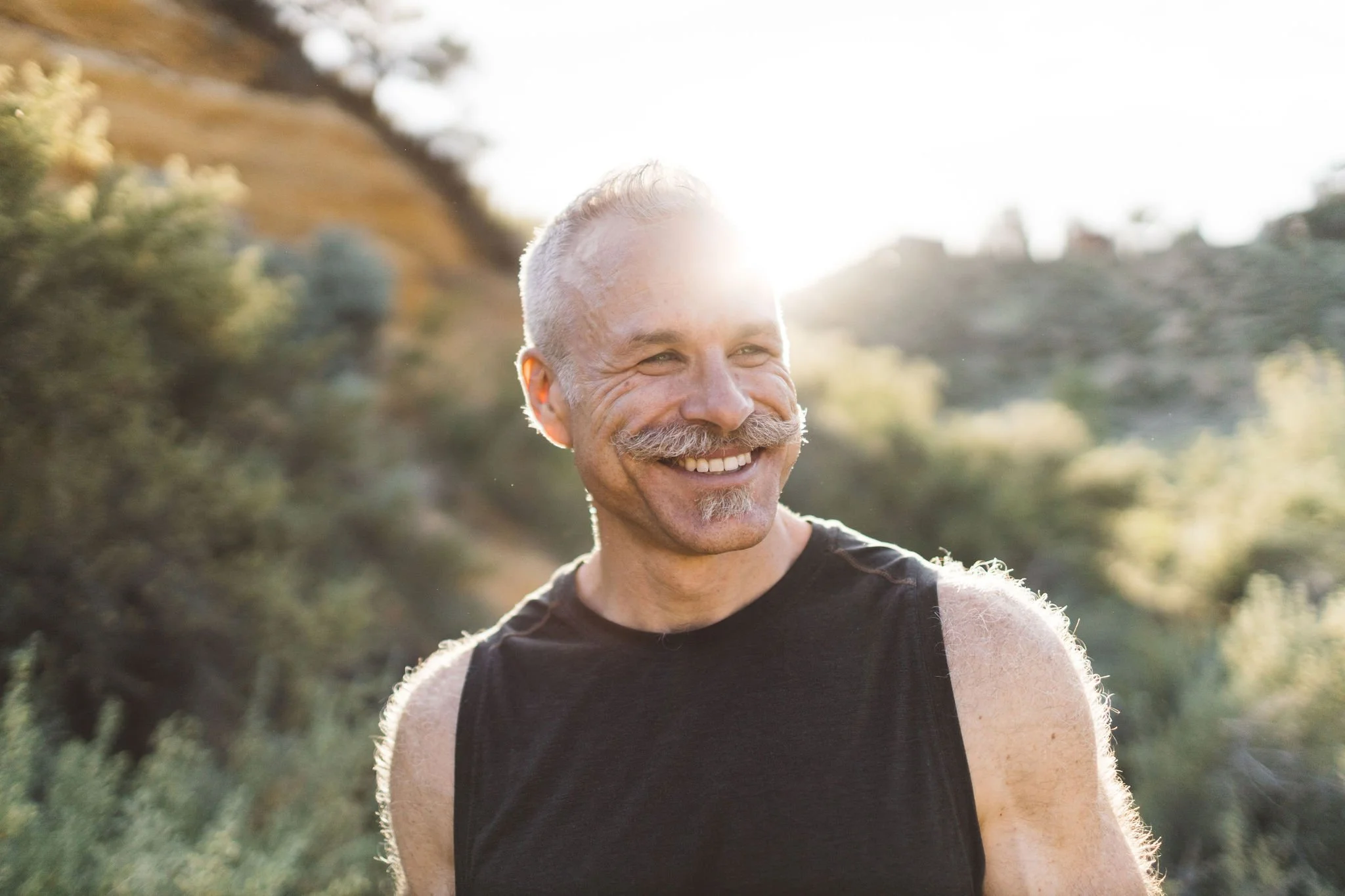 A smiling older man with gray hair and a gray mustache, wearing a sleeveless black shirt, outdoors with a blurred background of trees and hills, backlit by sunlight.