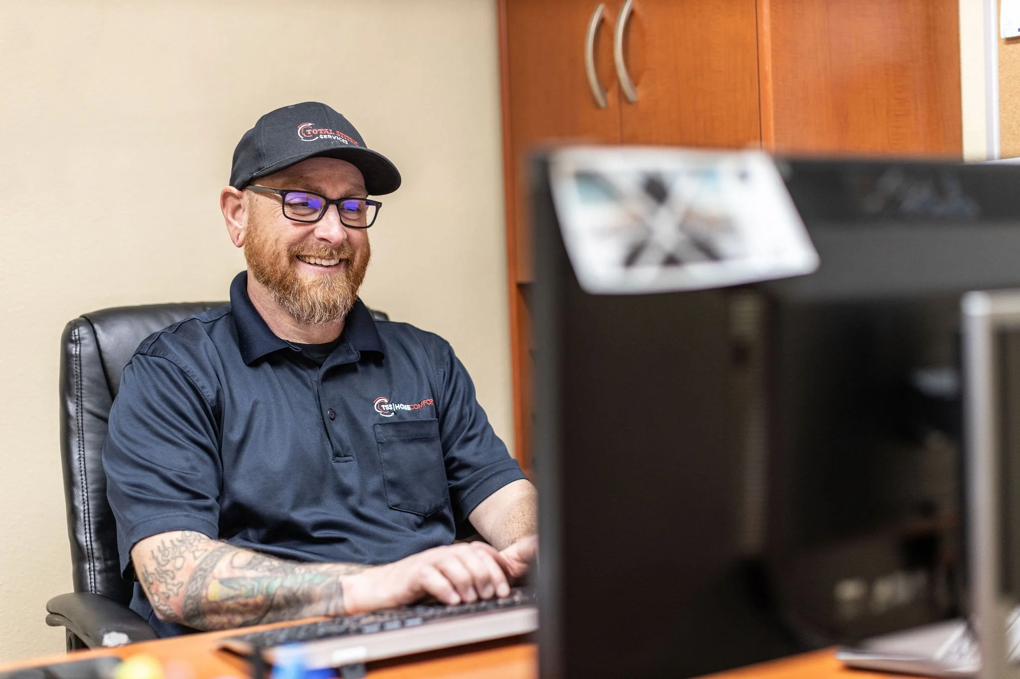 A smiling man with glasses and a beard, wearing a black cap and a black polo shirt, sitting at a desk and looking at a computer monitor in an office.