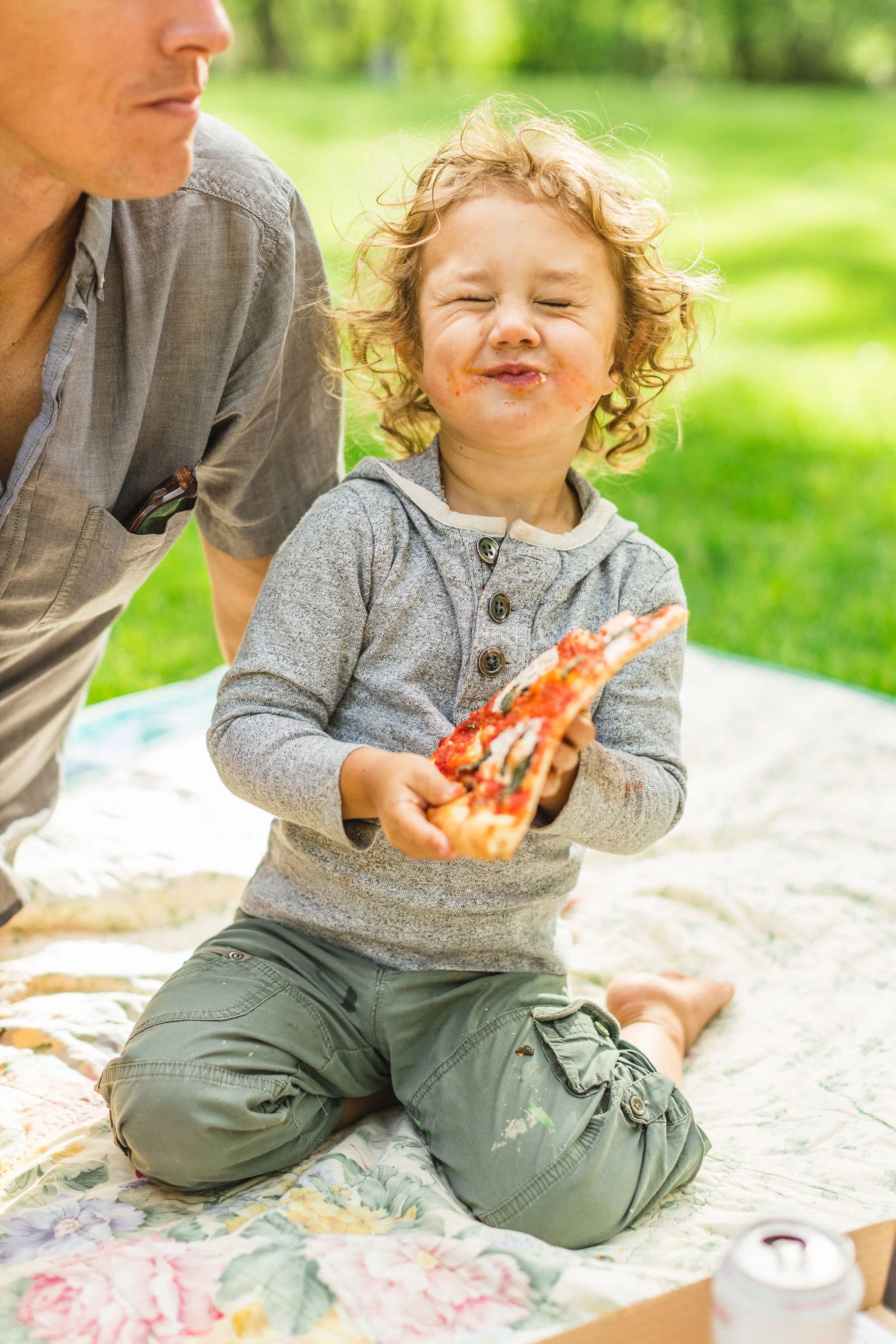 Young child with curly blonde hair, kneeling on a floral blanket outdoors, holding a slice of pizza, with a parent standing next to them, all in a green park setting.