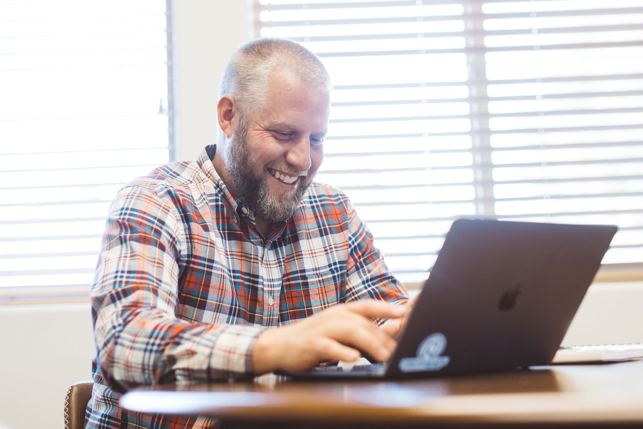 Business owner with a beard smiling during a branding photo session while using a laptop at a desk in front of window blinds in Boise, Idaho.