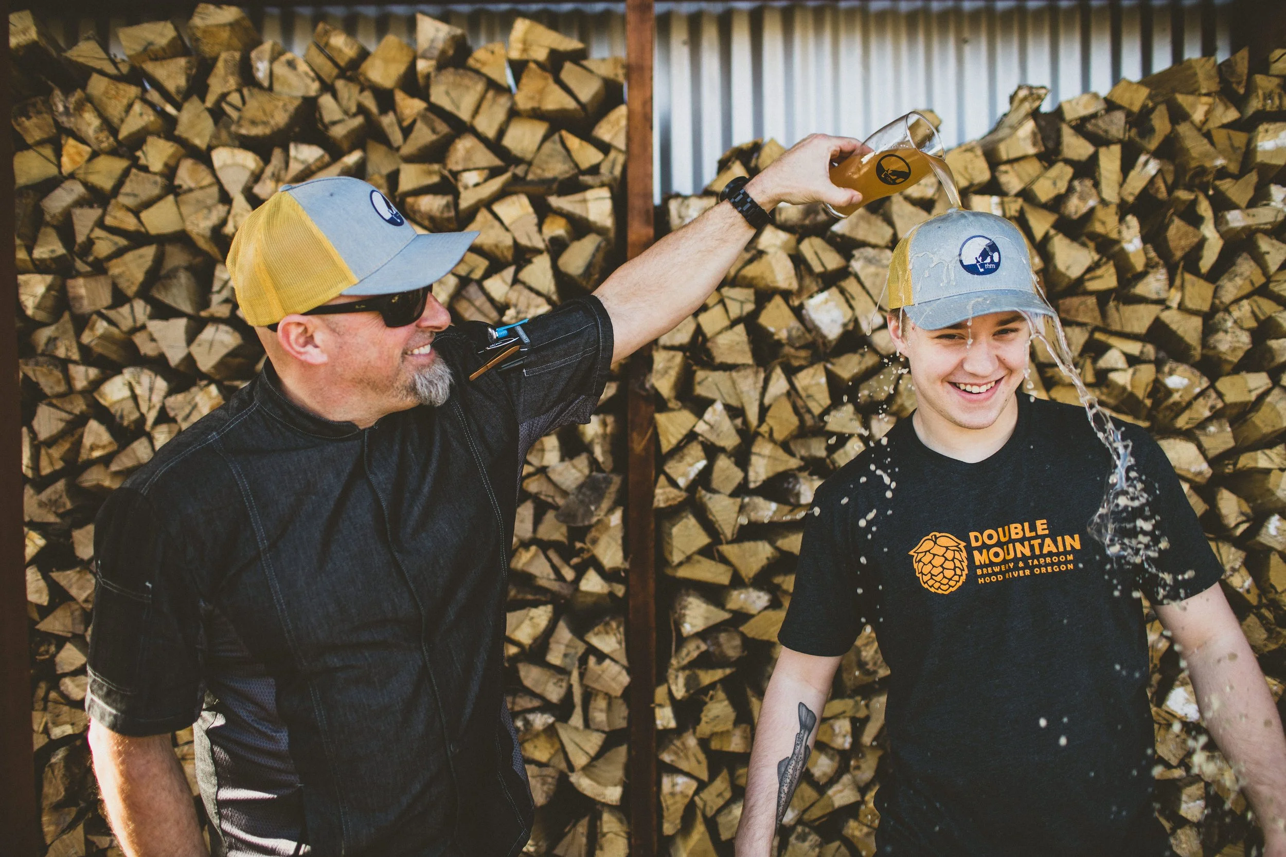 Two men celebrating with beer during a branding photoshoot.  The man is pouring a beer over the other man's head, and they are both smiling. They are wearing hats and sunglasses, and the background shows a large stack of chopped firewood.