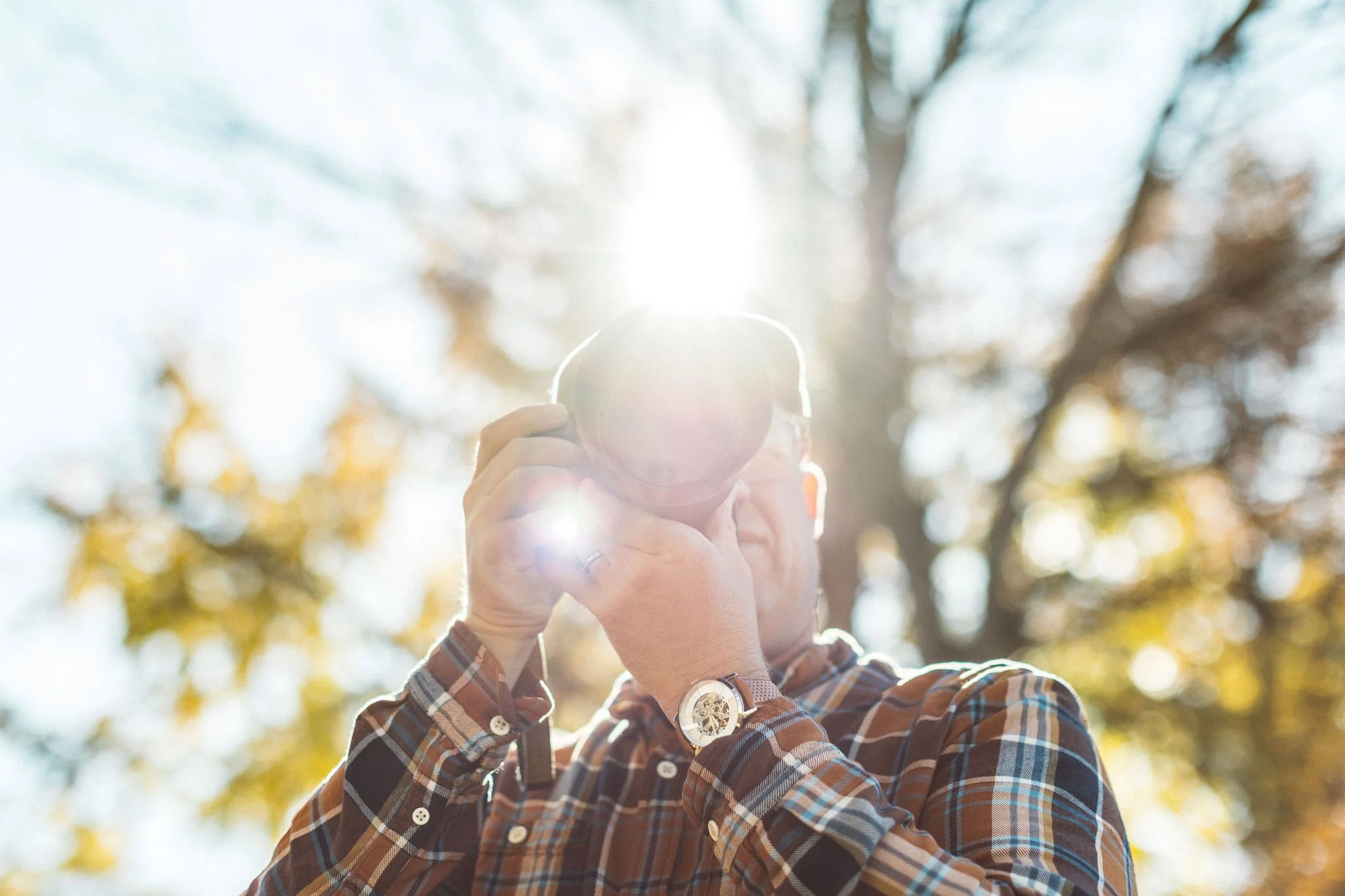 Bryan Rupp holding a camera outdoors with sunlight shining behind him, wearing a plaid shirt and a watch in Boise idaho.