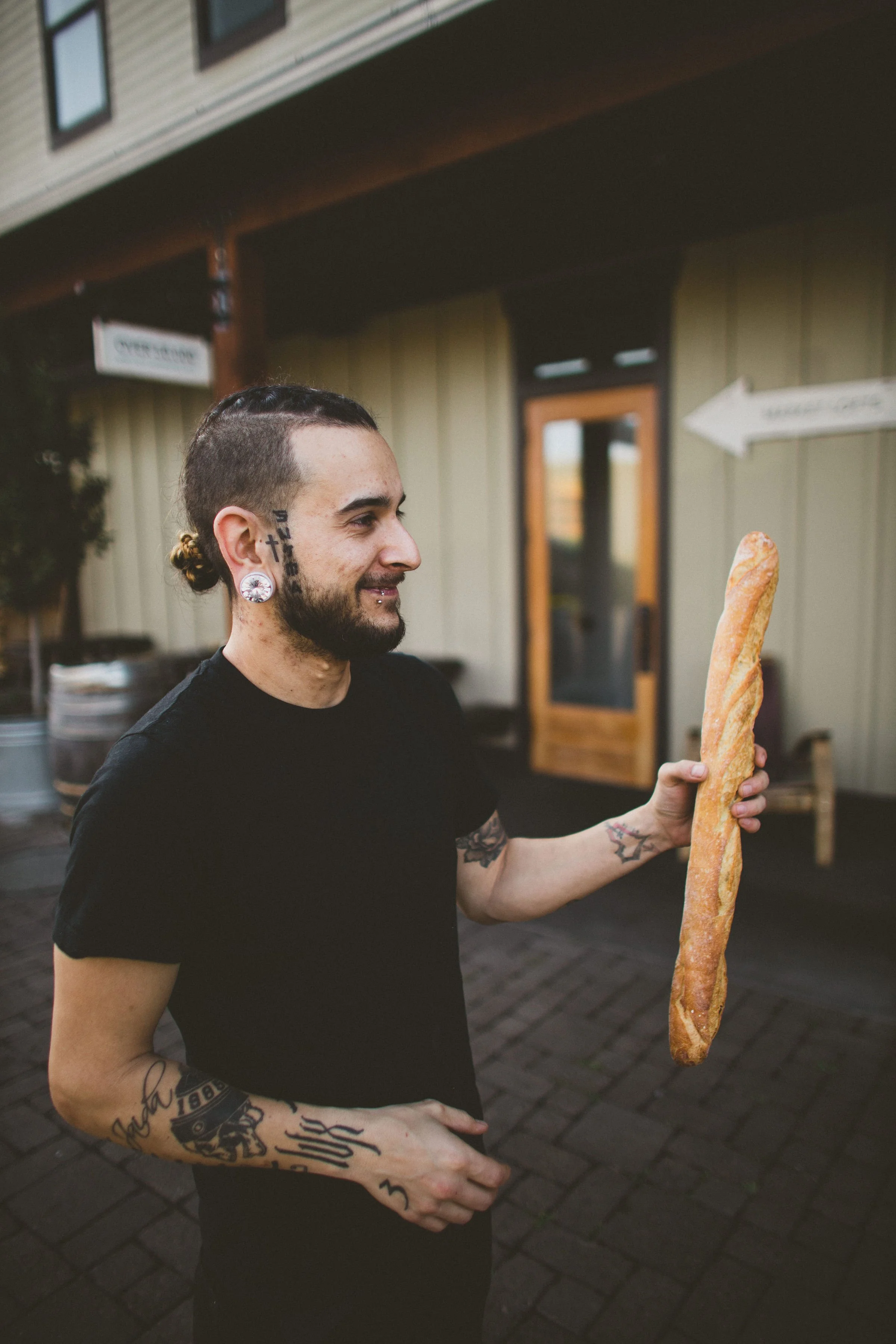 A man with tattoos and piercings holding a large baguette outside a building during a branding photoshoot in Dundee, Oregon.