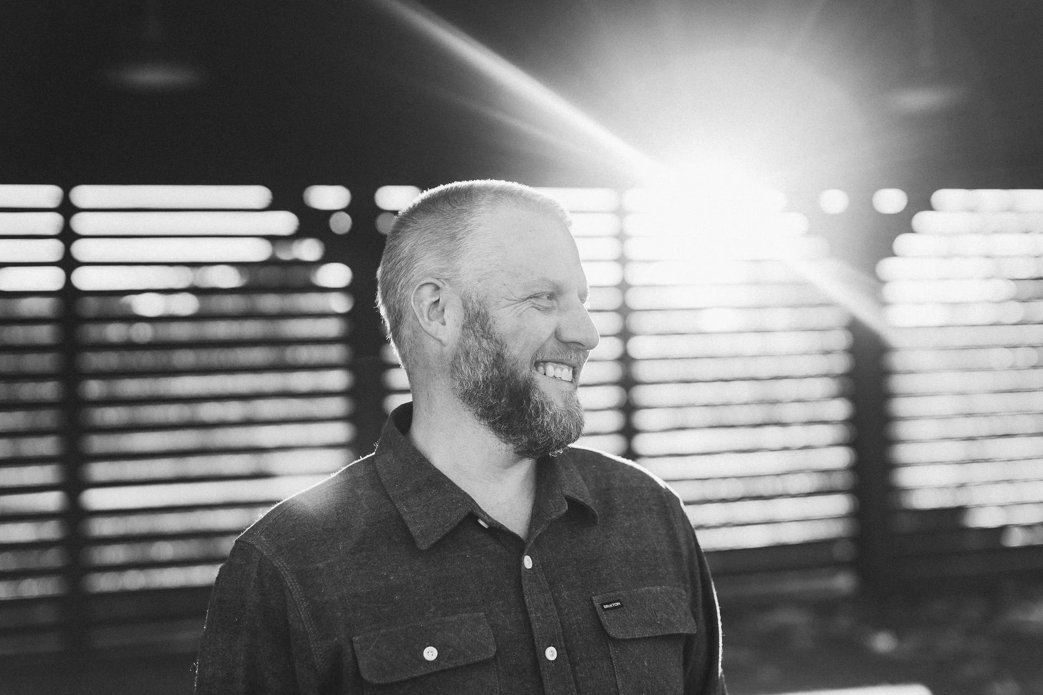 A smiling business owner with a beard looking to the side during a branding photoshoot, in front of a background at a park in Boise with horizontal light bars and bright sunlight creating a backlit effect.