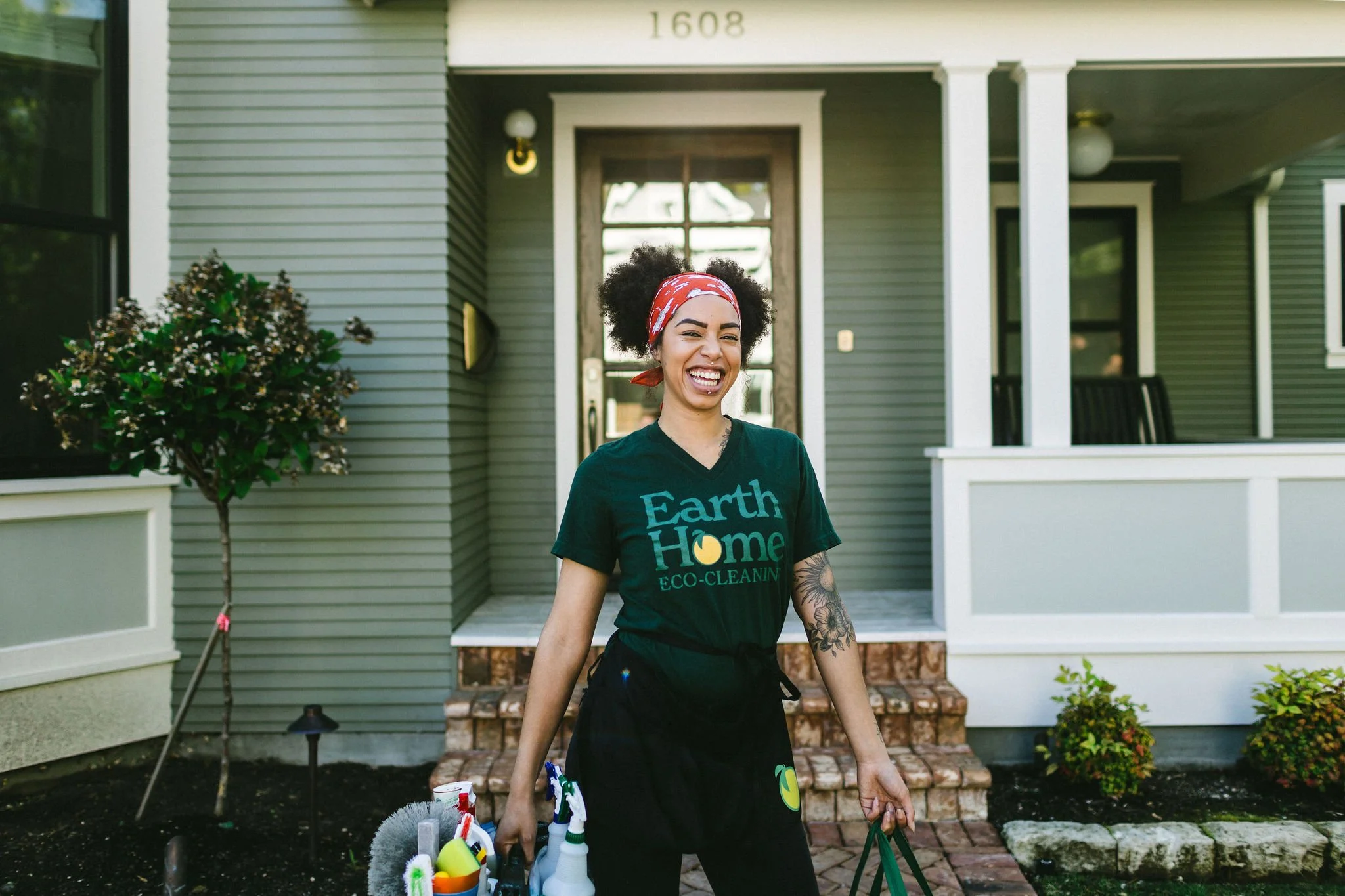 Branding photo of a woman house cleaner entrepreneur standing in front of a home smiling and holding cleaning supplies during a branding photoshoot in Boise, Idaho.
