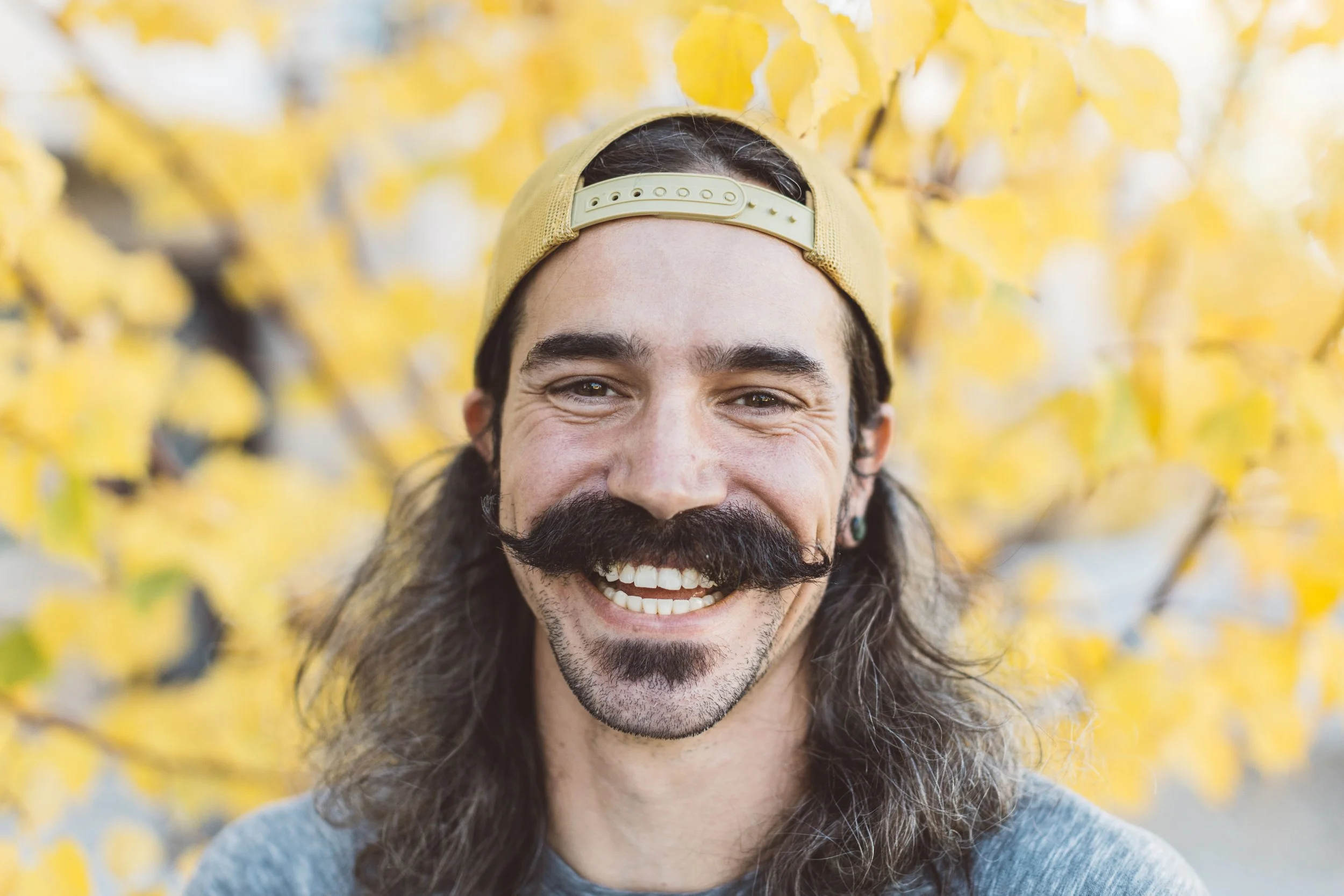 An entrepreneur with long hair and a mustache smiling outdoors with yellow autumn leaves in the background during a branding photoshoot in Boise, Idaho.