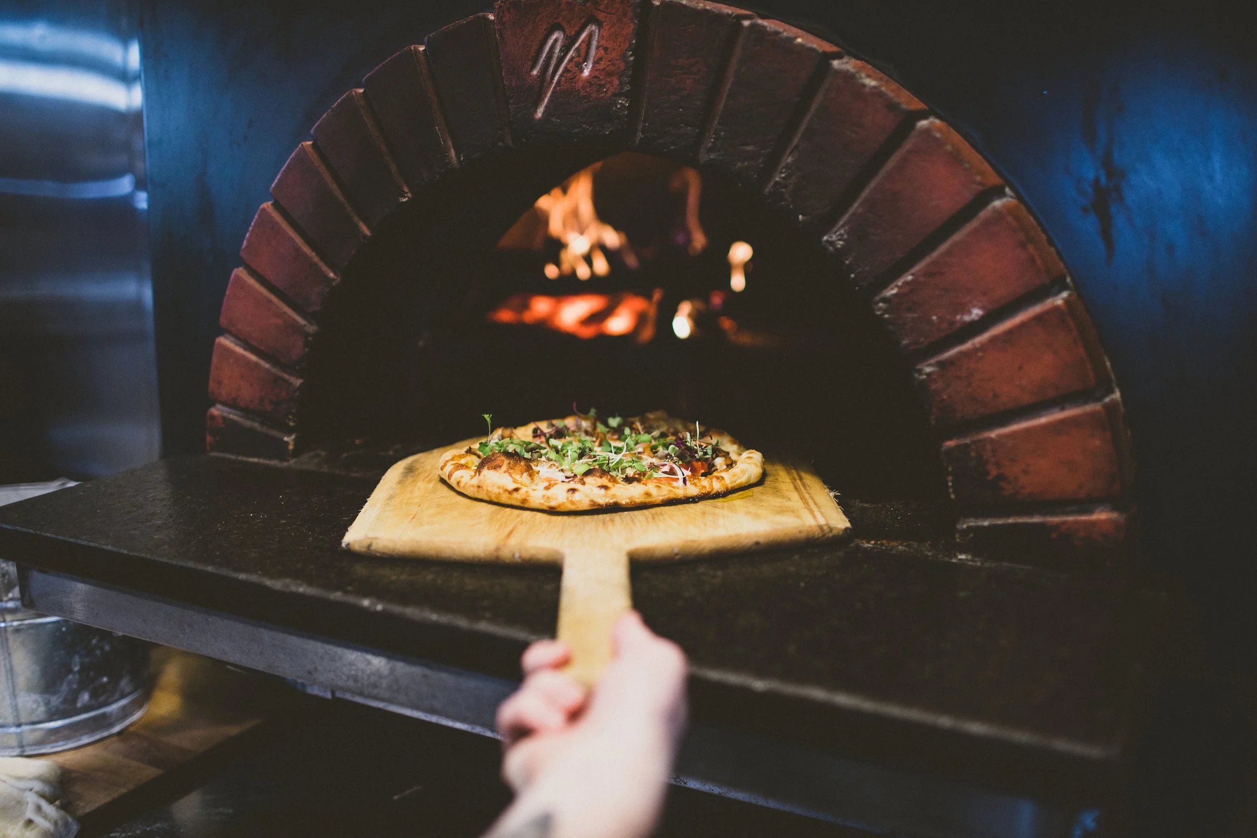 Person placing a pizza with greens onto a wooden paddle into a brick wood-fired oven for baking during a branding photoshoot in Dundee, Oregon.