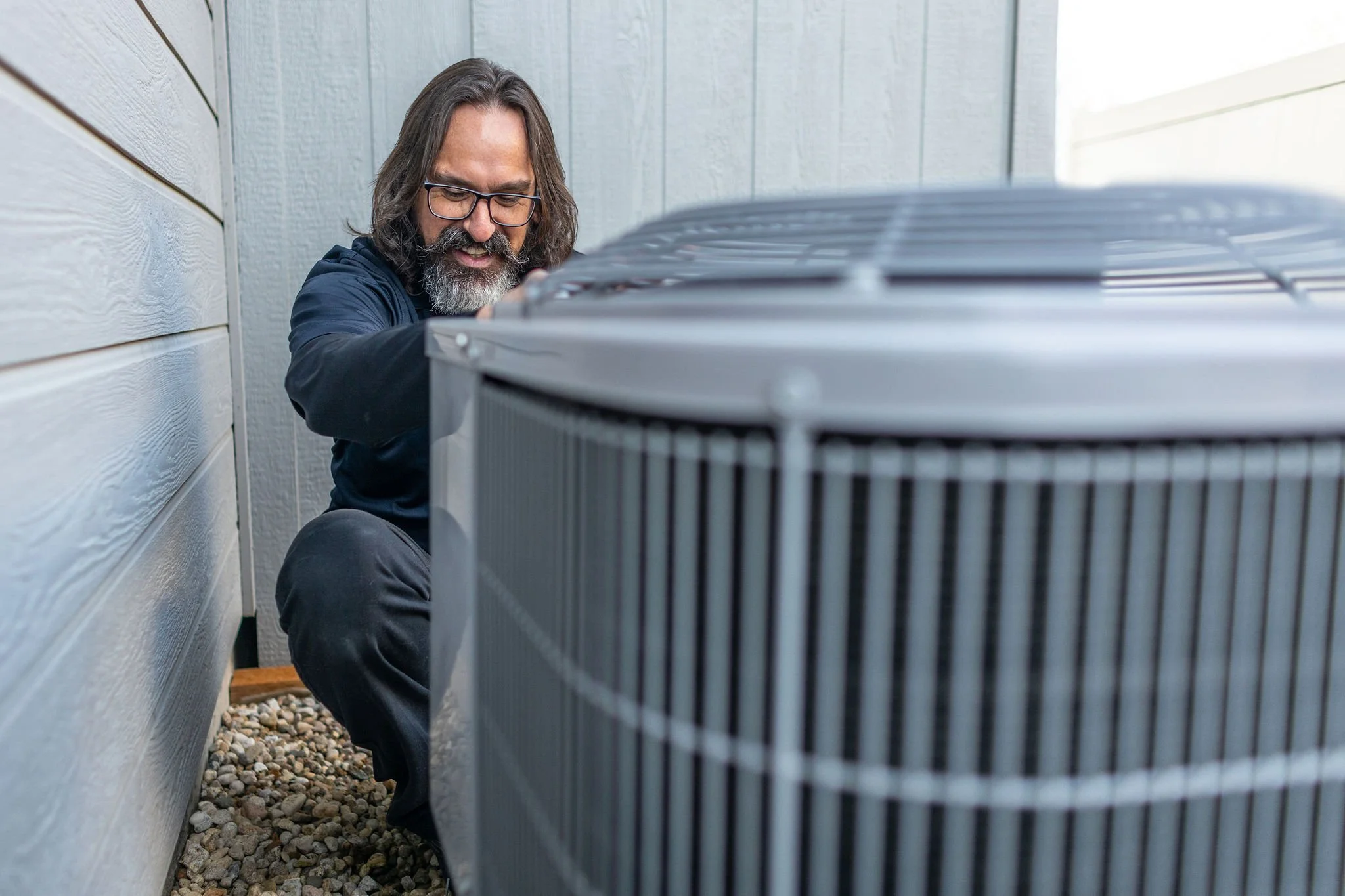 A man with long hair, glasses, and a beard repairs an outdoor air conditioning unit beside a house with light-colored siding.
