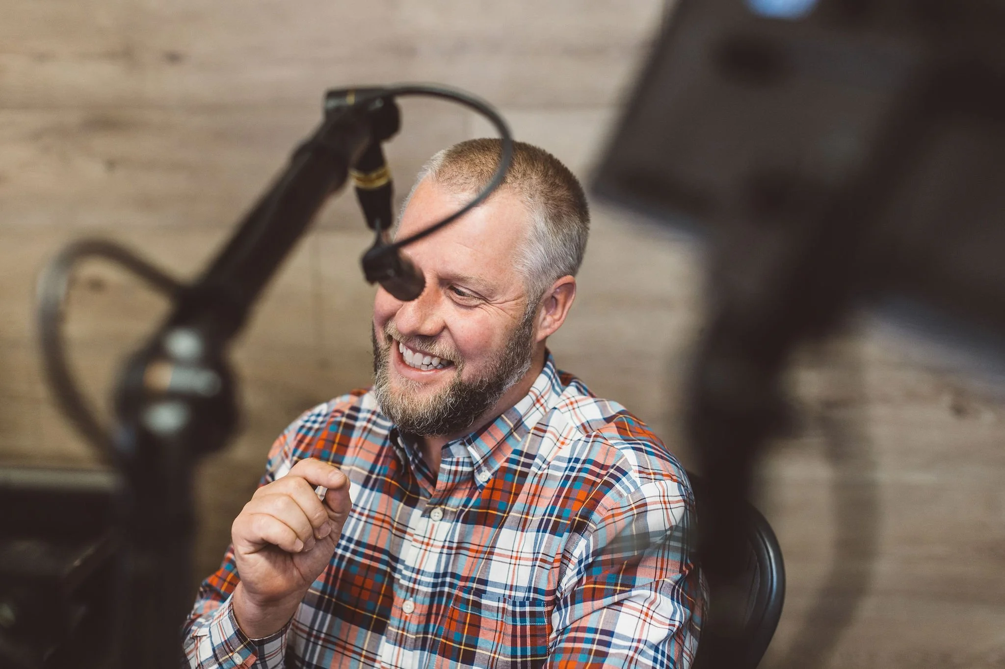 A smiling entrepreneur with a beard and short hair, wearing a plaid shirt, speaking into a microphone during an interview or podcast recording in a studio in Boise Idaho during a branding photoshoot.