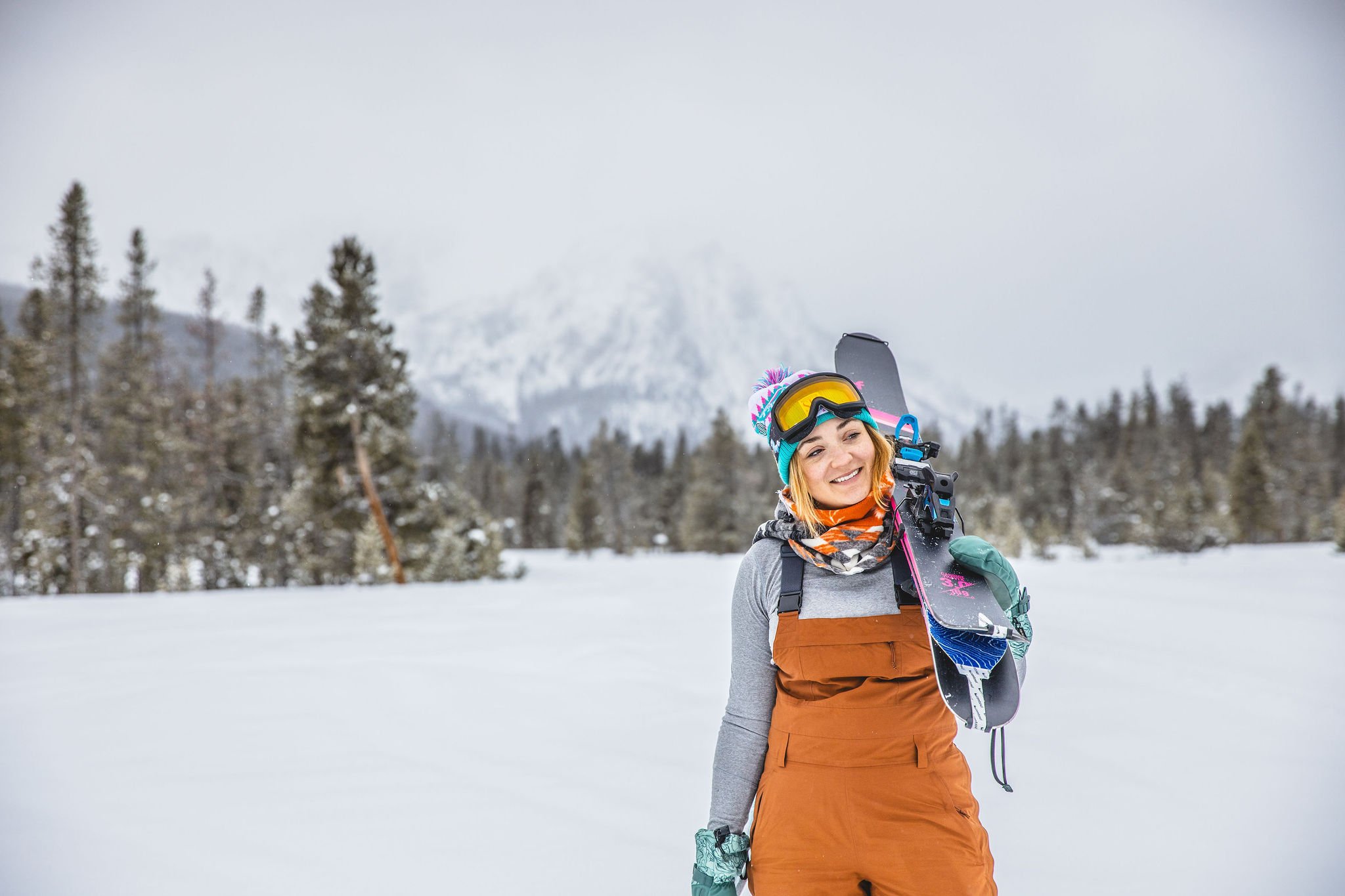 A woman standing in a snowy landscape holding a snowboard over her shoulder, wearing winter gear and smiling, with pine trees and snow-covered mountains in the background.