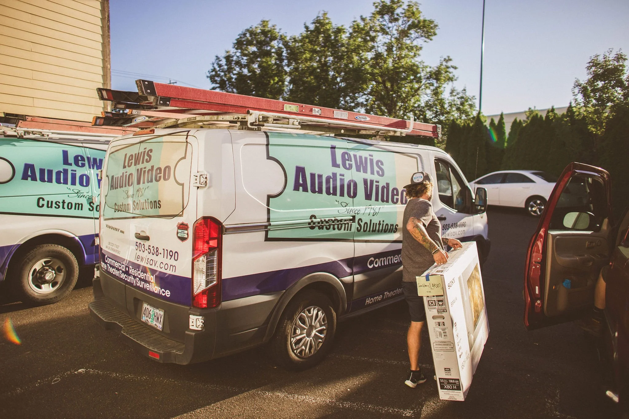 A man unloading a boxed television from the back of a Lewis Audio Video service van, with ladders on top, at a parking lot during daytime, during a branding photoshoot in Newberg, Oregon.