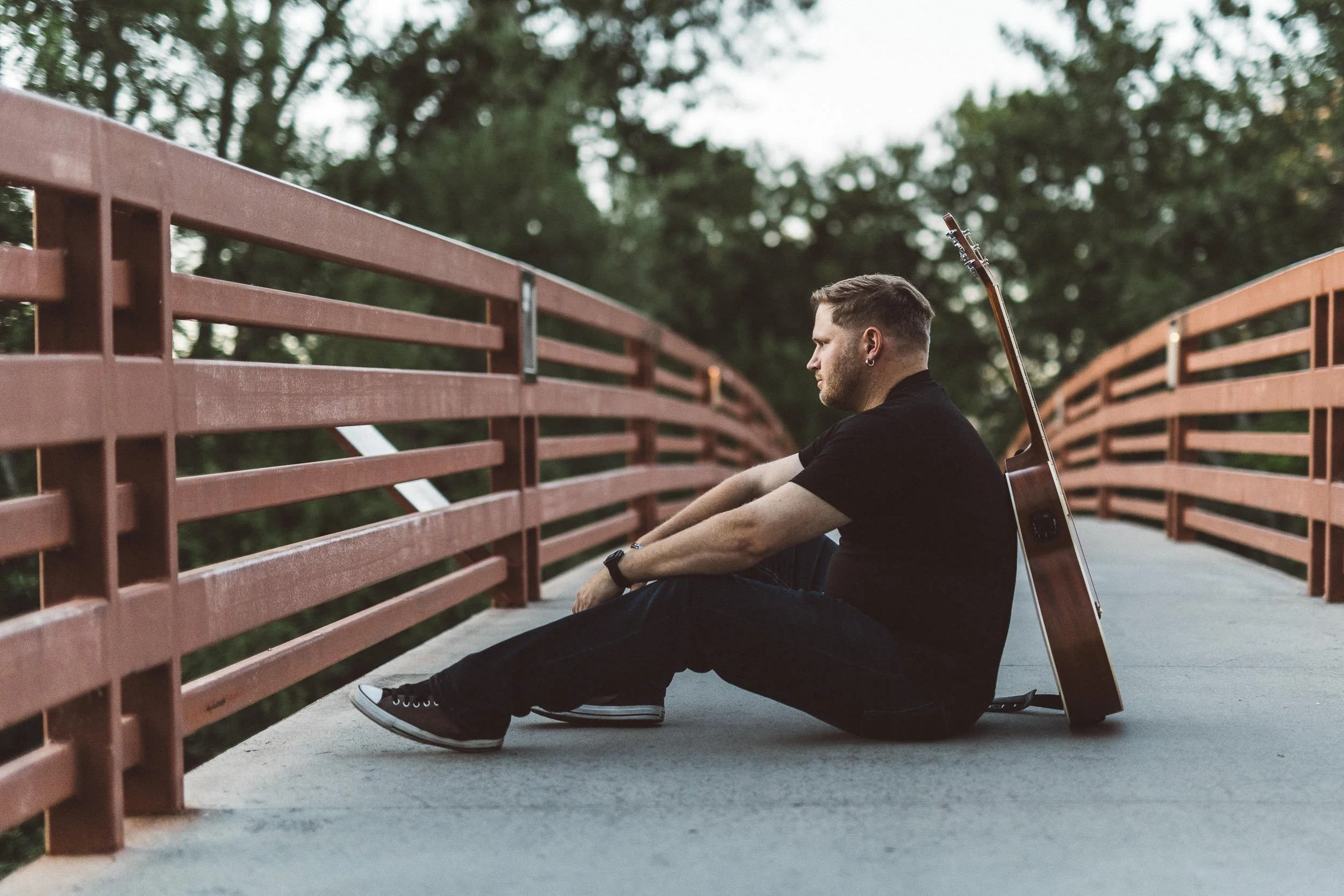 Man sitting on bridge with guitar leaning against his back during a branding photoshoot at Julia Davis Park at sunset.