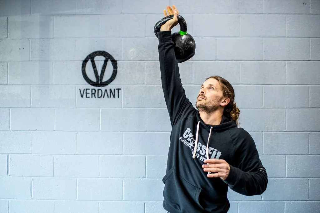 A man lifting a kettlebell above his head in a gym with a gray brick wall behind him