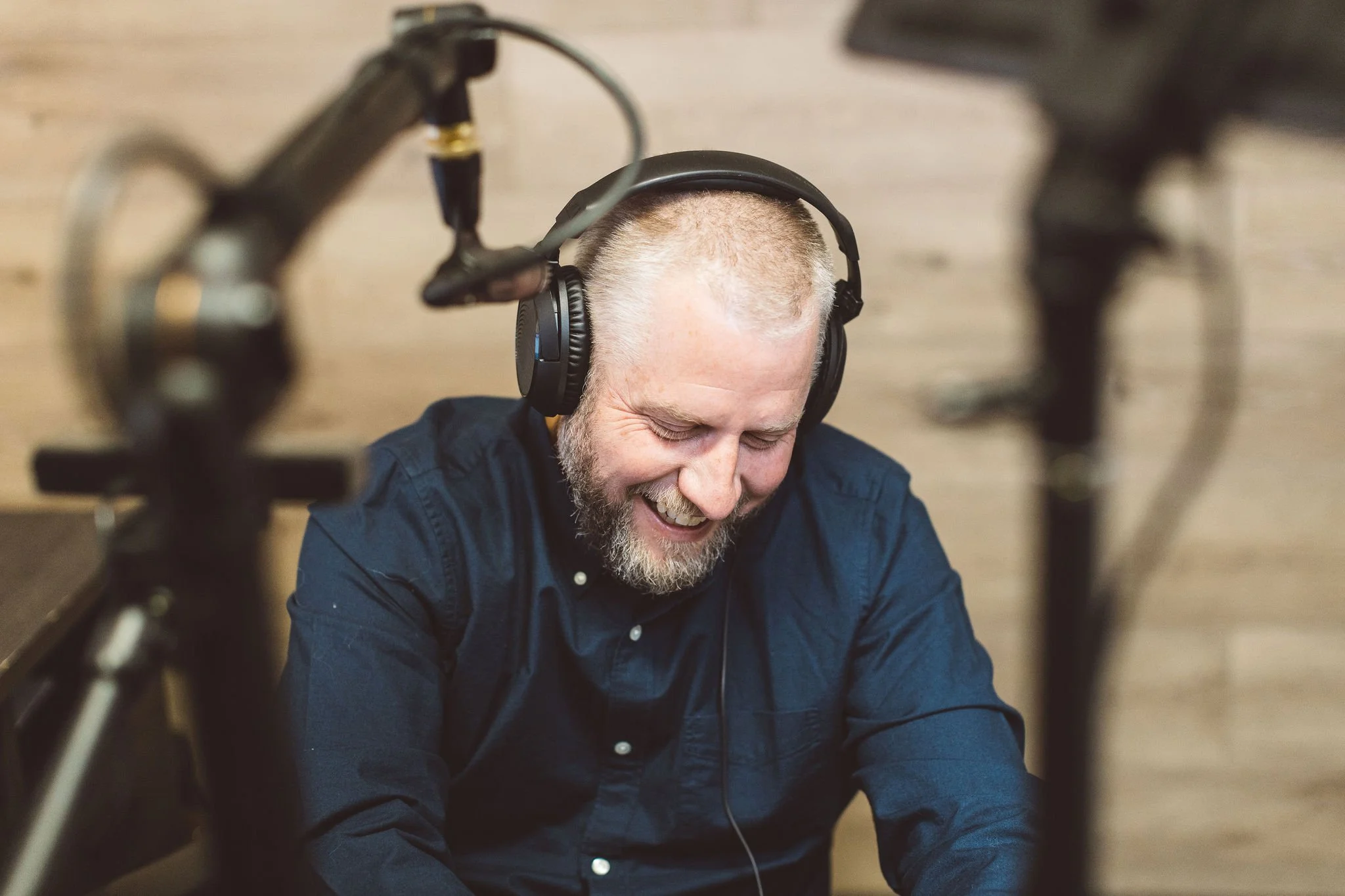 A man with a beard and short hair, smiling and wearing headphones, sitting in front of microphone equipment which is slightly out of focus in the foreground.