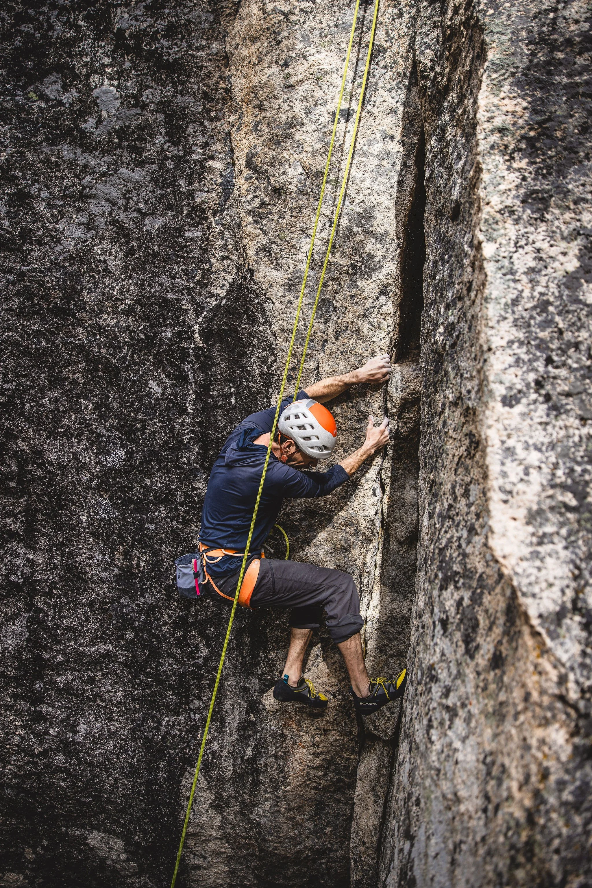 A male rock climber wearing a helmet and harness is scaling a rock face, with a climbing rope secured to his harness.