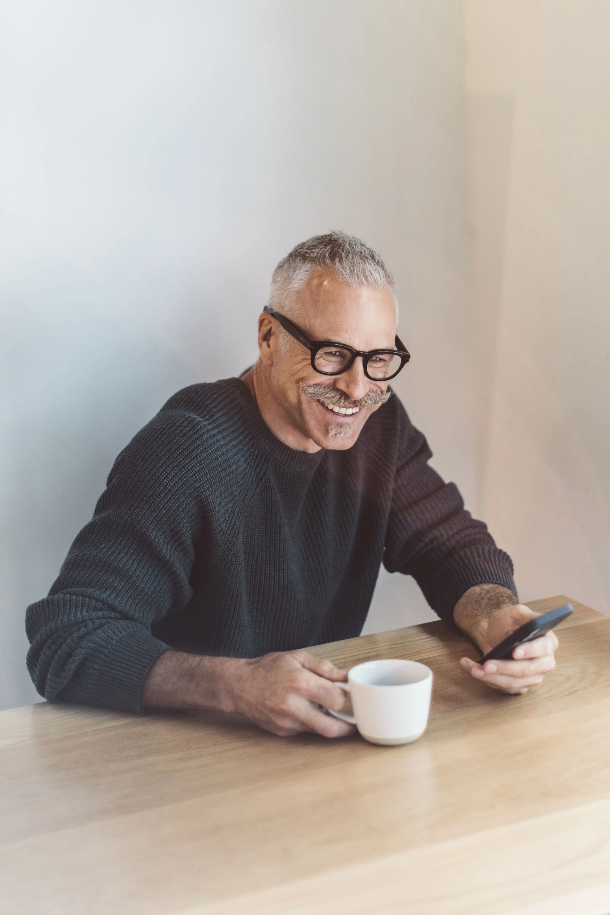 Smiling older man with gray hair and glasses sitting at a wooden table, holding a smartphone in one hand and a coffee cup in the other.