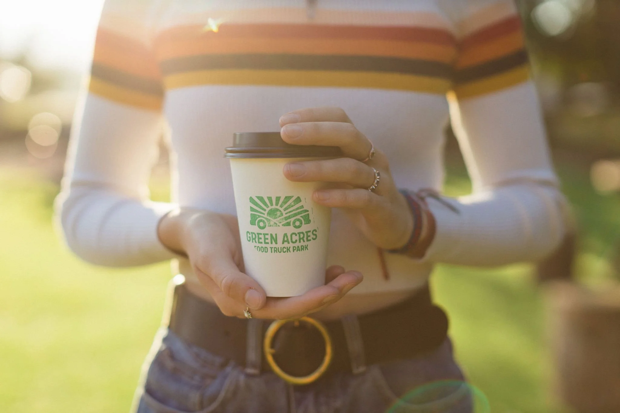 Person holding a to-go coffee cup with Green Acres Food Truck Park logo, wearing a striped shirt and jeans, outdoors with sunlight and greenery in the background.