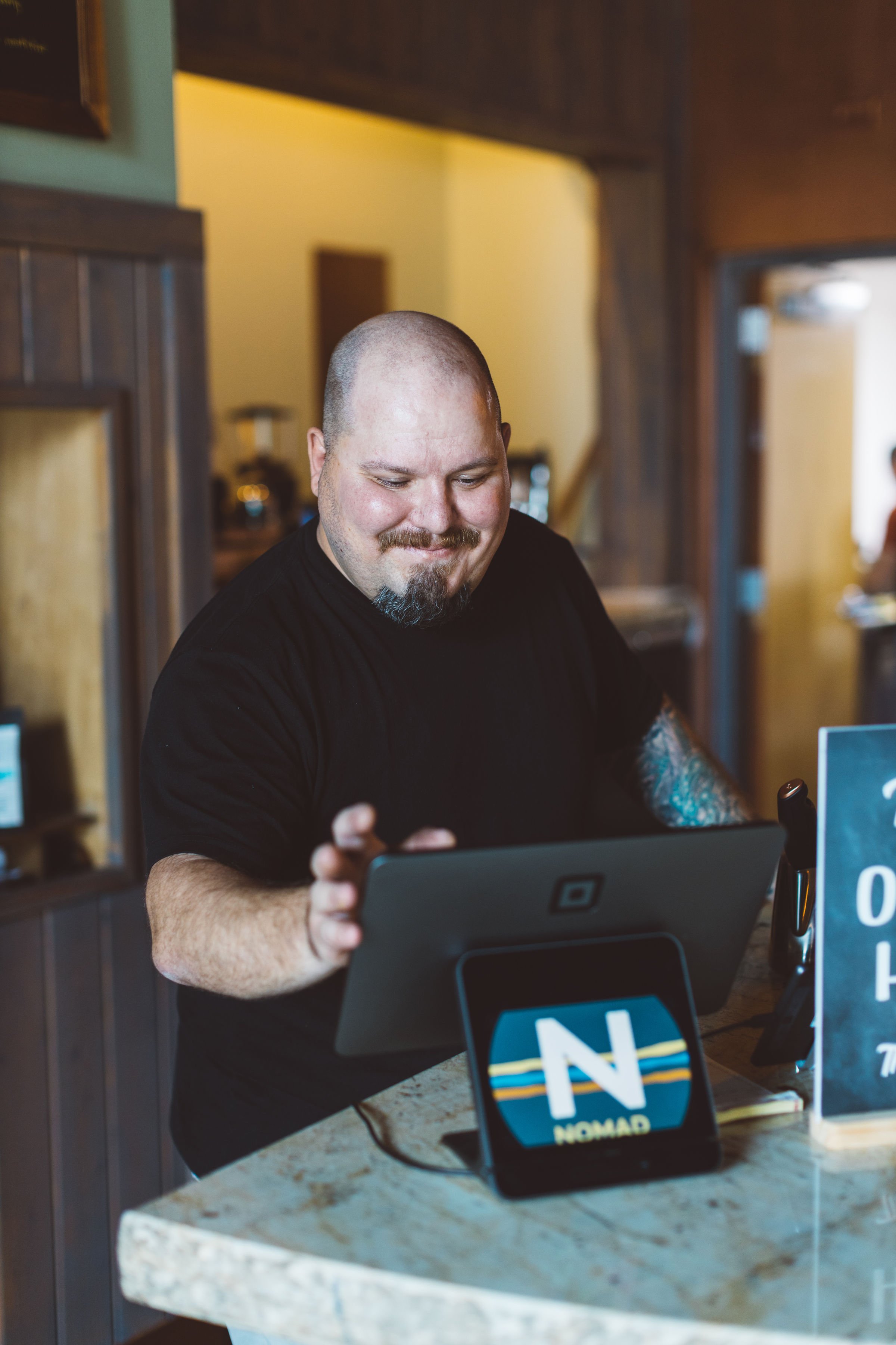 A business owner with a shaved head, beard, and tattoos on his right arm is smiling while using a tablet at a restaurant during a branding photoshoot in Boise, Idaho.