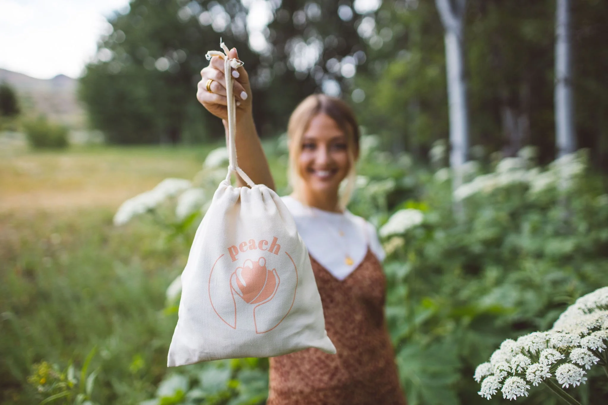 A woman business owner holds a small carry bag with a peach logo and the word 'peach' printed on it, outdoors in a green park or garden during a branding photoshoot in Boise, Idaho.