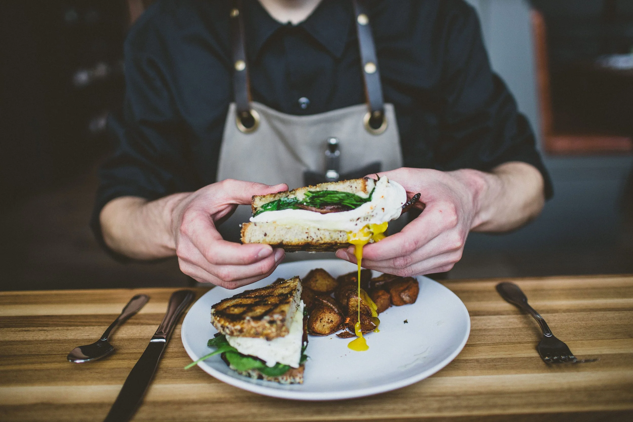 A person wearing a black shirt and apron holding a sandwich with egg, spinach, and other ingredients.