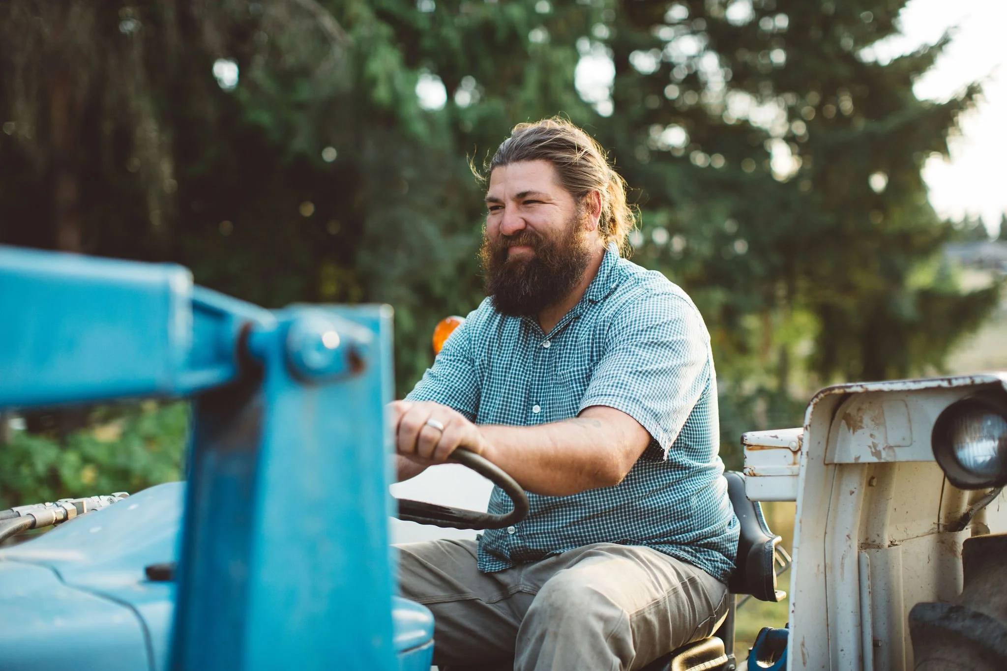 A man with a beard and long hair, wearing a blue checkered shirt, is sitting on a tractor outdoors, smiling slightly with trees in the background.