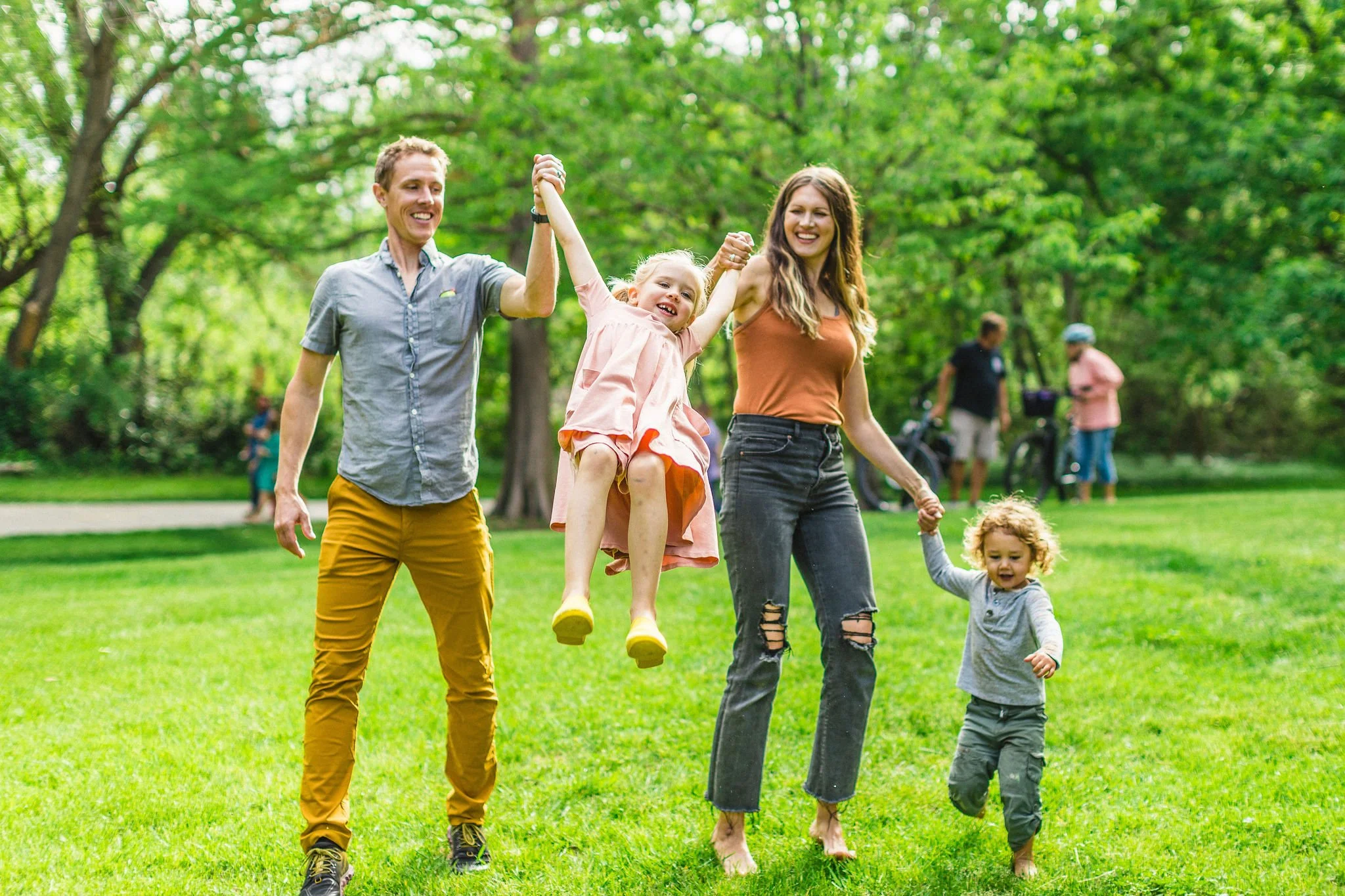 A happy family of four, two adults and two children, enjoying time outdoors in a park. The mother is holding one child's hand while the father lifts the other child in the air, all smiling and playing on a grassy area surrounded by trees.