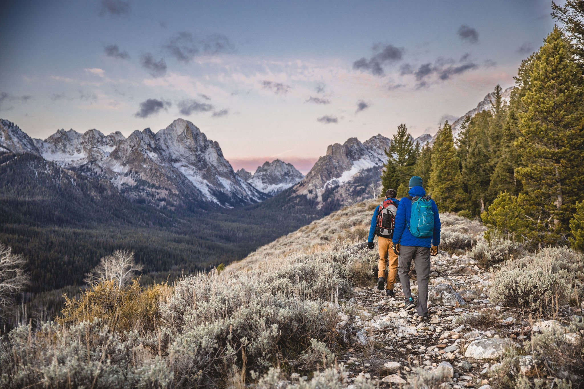 Three hikers walking on a trail in a mountain landscape with snow-capped peaks, green trees, and sunrise or sunset sky.