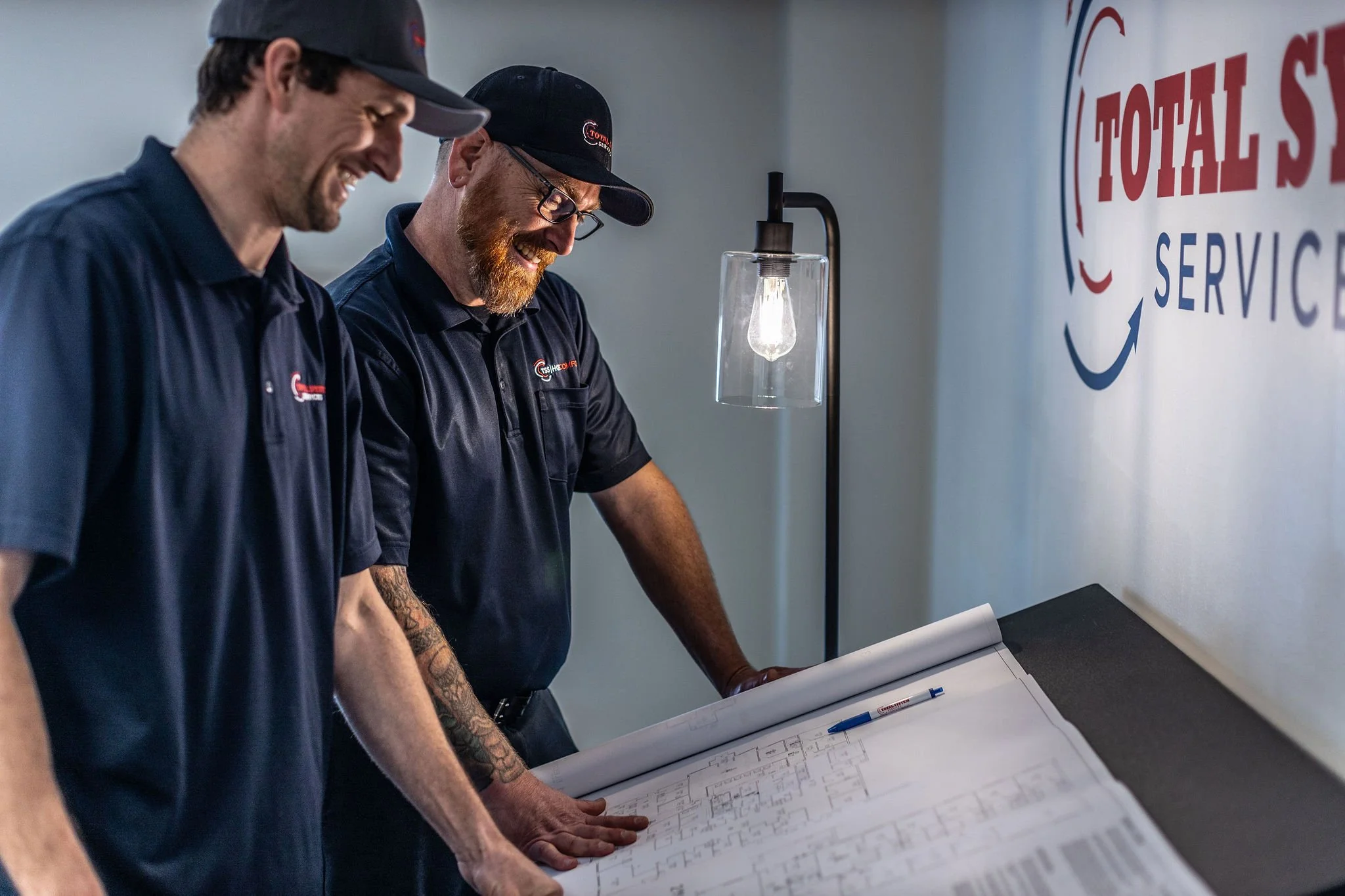 Two men in matching navy blue shirts and caps stand over a large blueprint on a table, smiling and discussing. Behind them is a wall with a logo that reads 'TOTAL SERVICE' and a standing lamp with an exposed bulb.