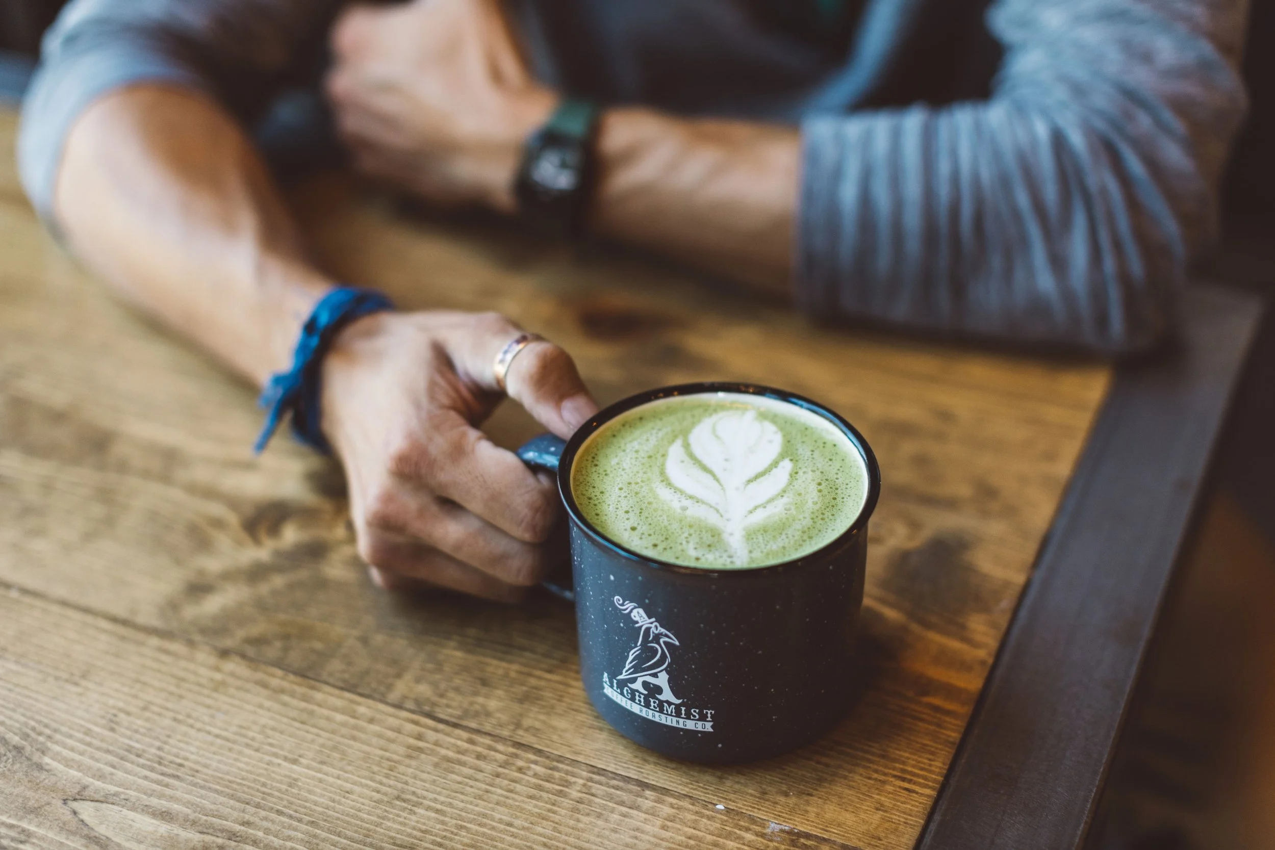 Person resting on a wooden table, holding a black mug with a green latte art design during a branding photoshoot in Boise, Idaho.