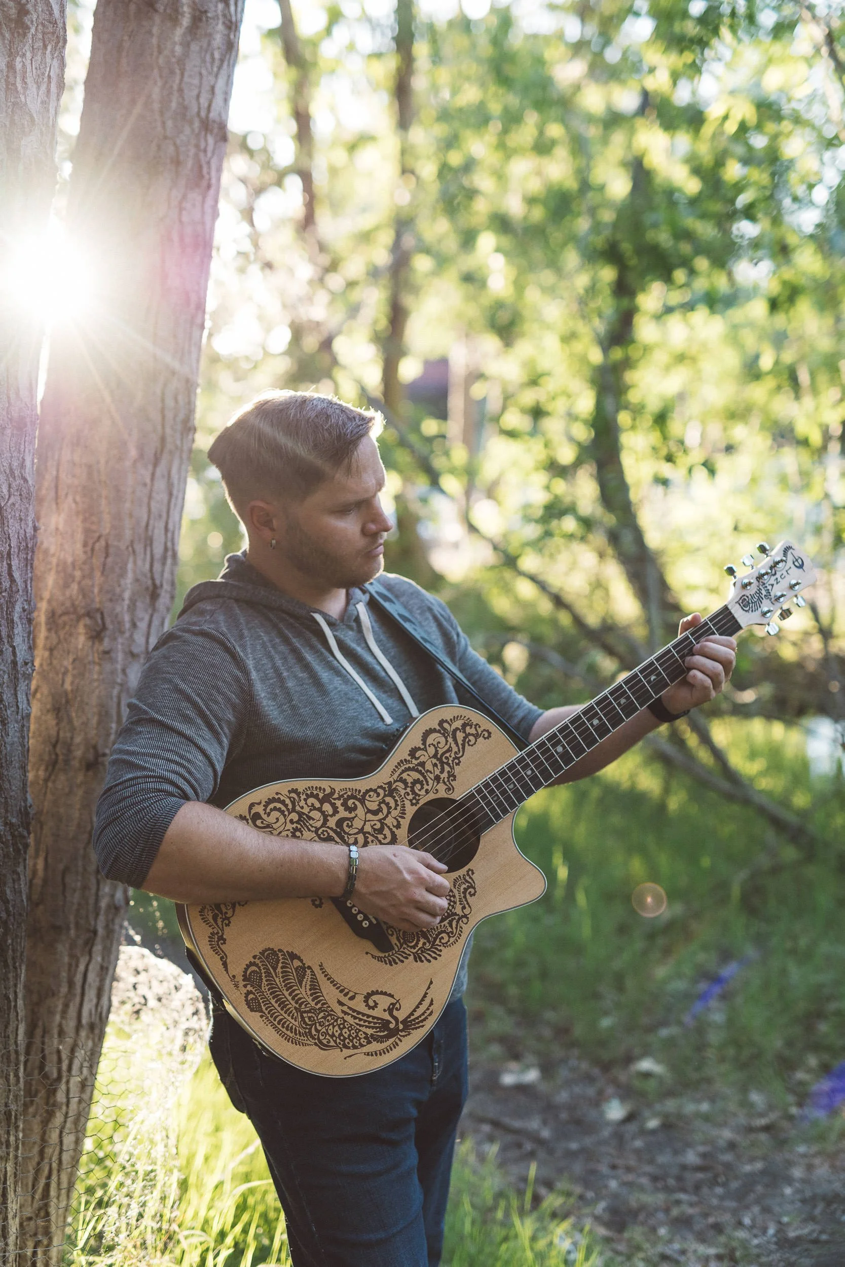 Man leaning against tree playing guitar on the Boise River Greenbelt near friendship bridge, during a branding photoshoot.