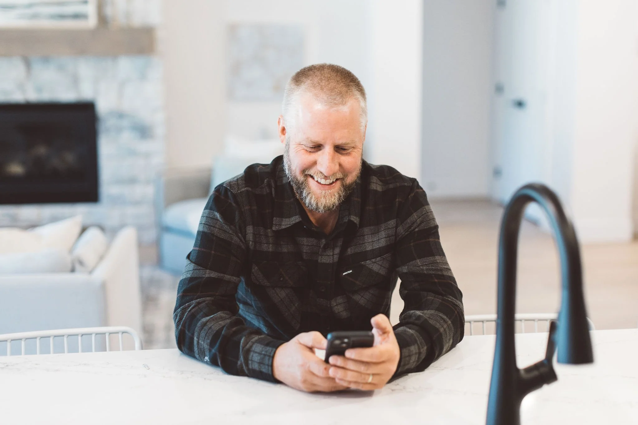 A smiling man with a beard and short hair, wearing a dark plaid shirt, looking at his smartphone while sitting at a white kitchen counter in a modern home.
