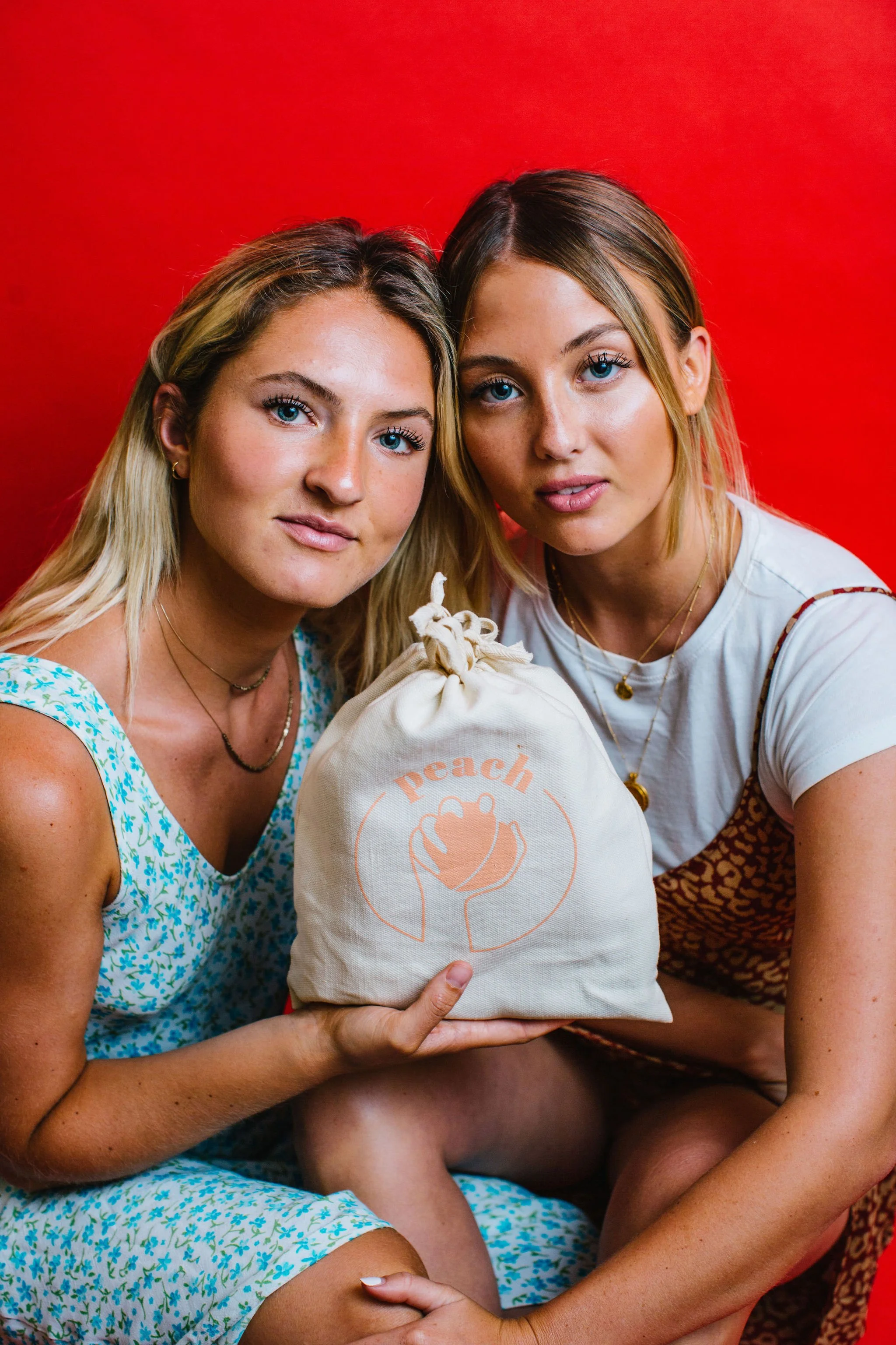 Two women with blonde hair and blue eyes holding a cloth bag with a peach logo and the word 'peach' against a red background during a studio. branding photoshoot in Boise, Idaho.