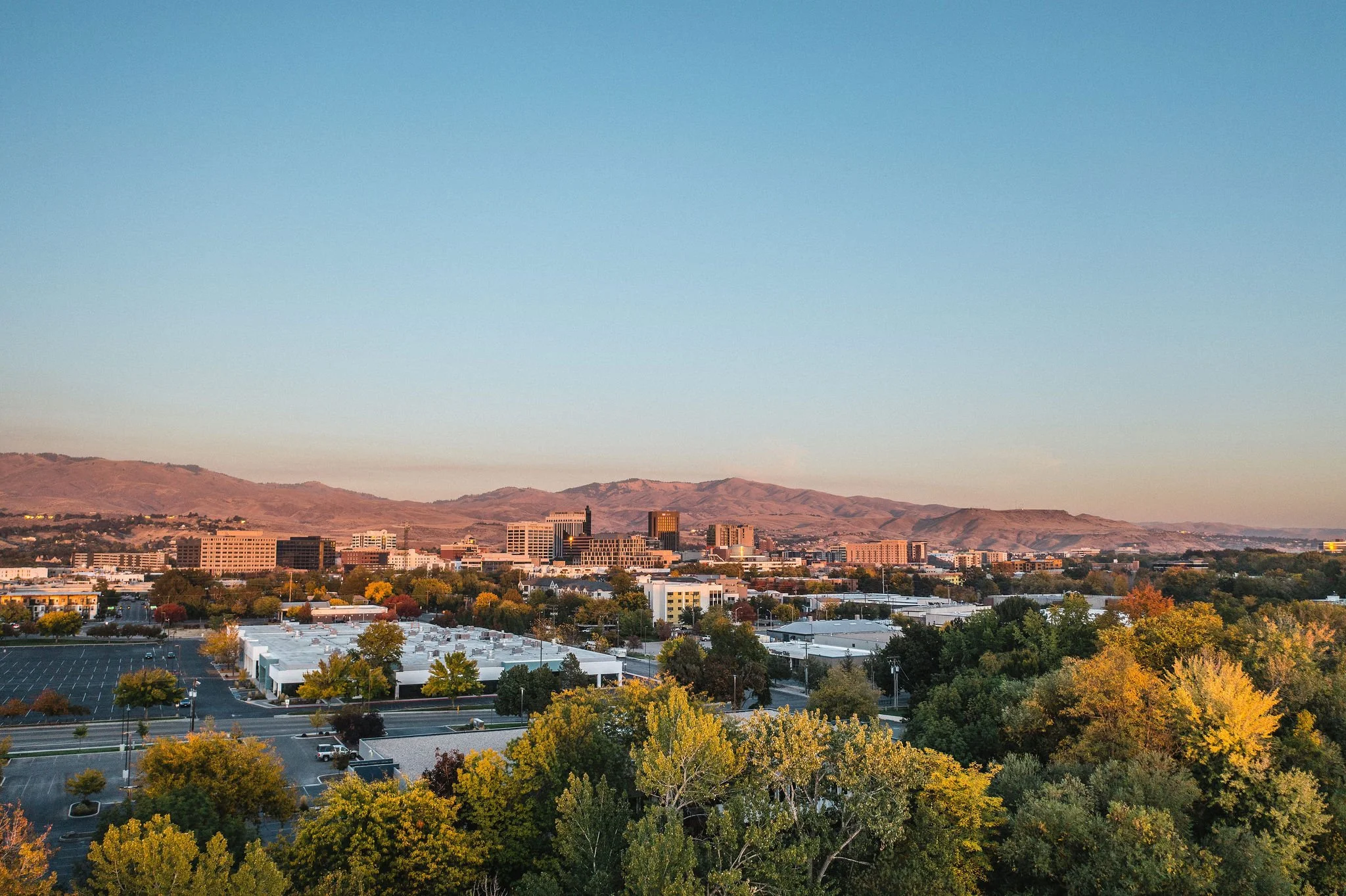 A cityscape with downtown buildings, surrounded by trees with fall foliage, hills in the background, and a parking lot in the foreground during sunset.