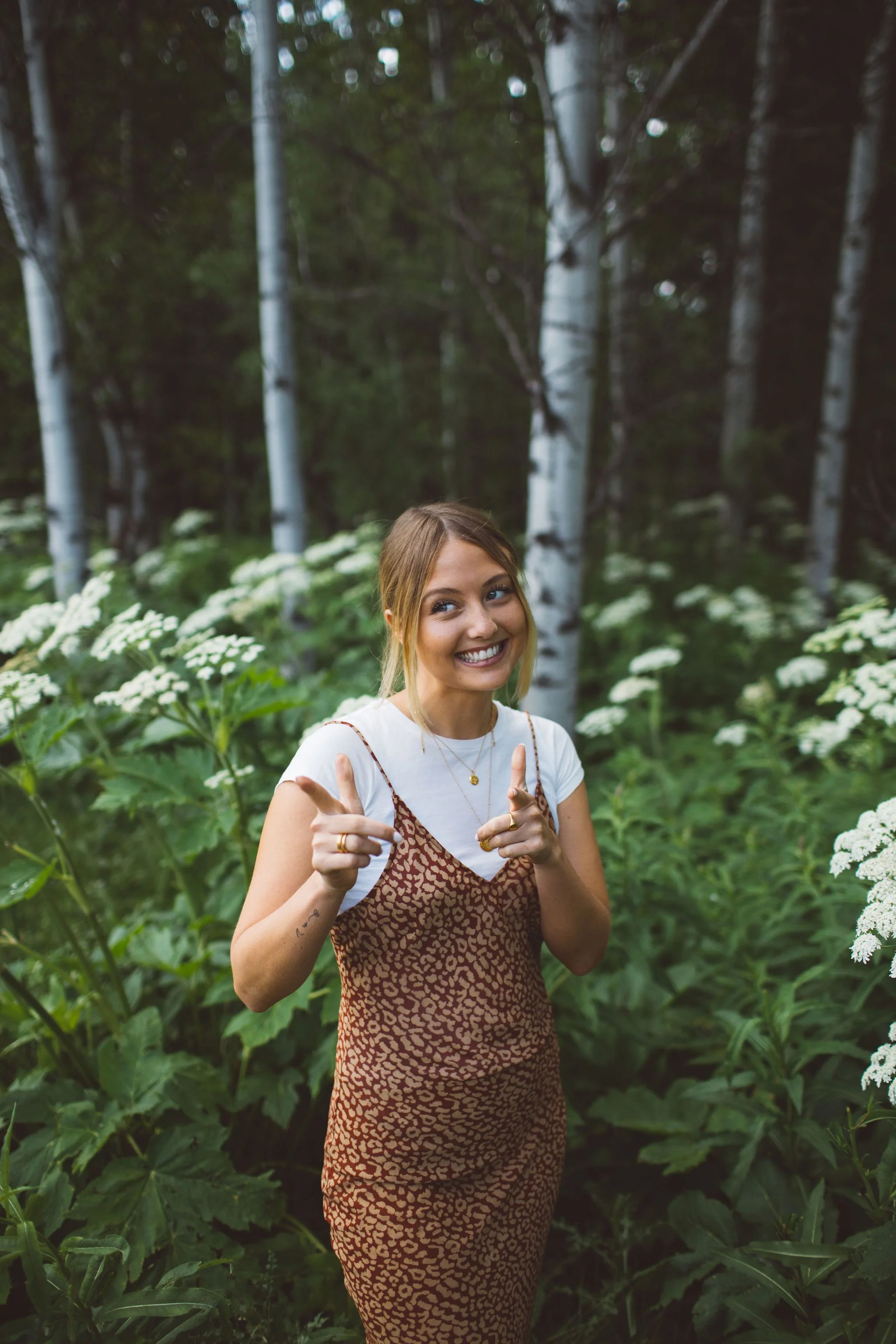 A young female business owner with blonde hair smiling and pointing with both hands while standing in a lush, green forest with white wildflowers and trees in the background, during a branding photoshoot in Sun Valley, Idaho.