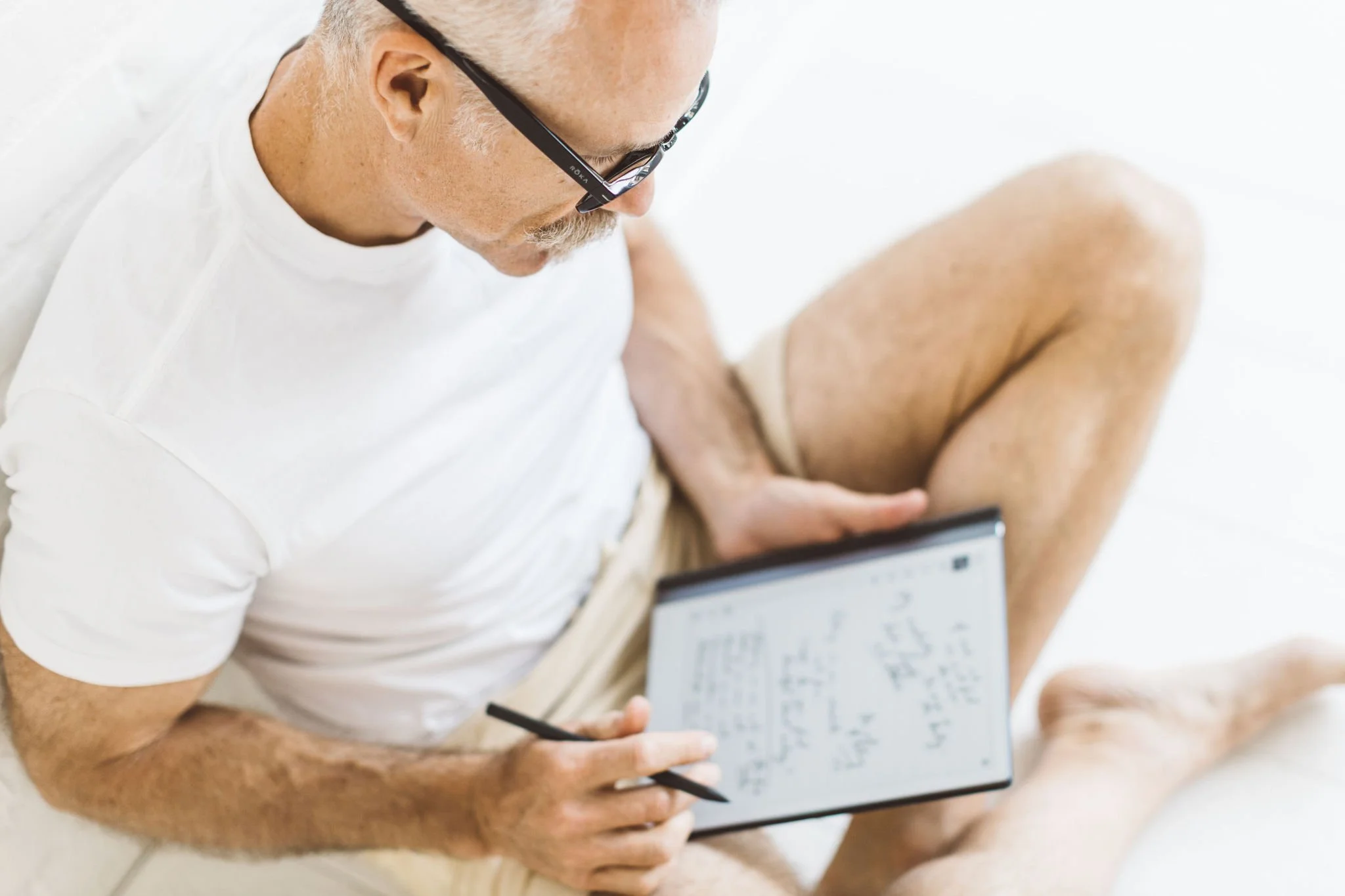 Older man with glasses wearing a white t-shirt, sitting on the floor with one knee up, holding a tablet and stylus, writing on the tablet.
