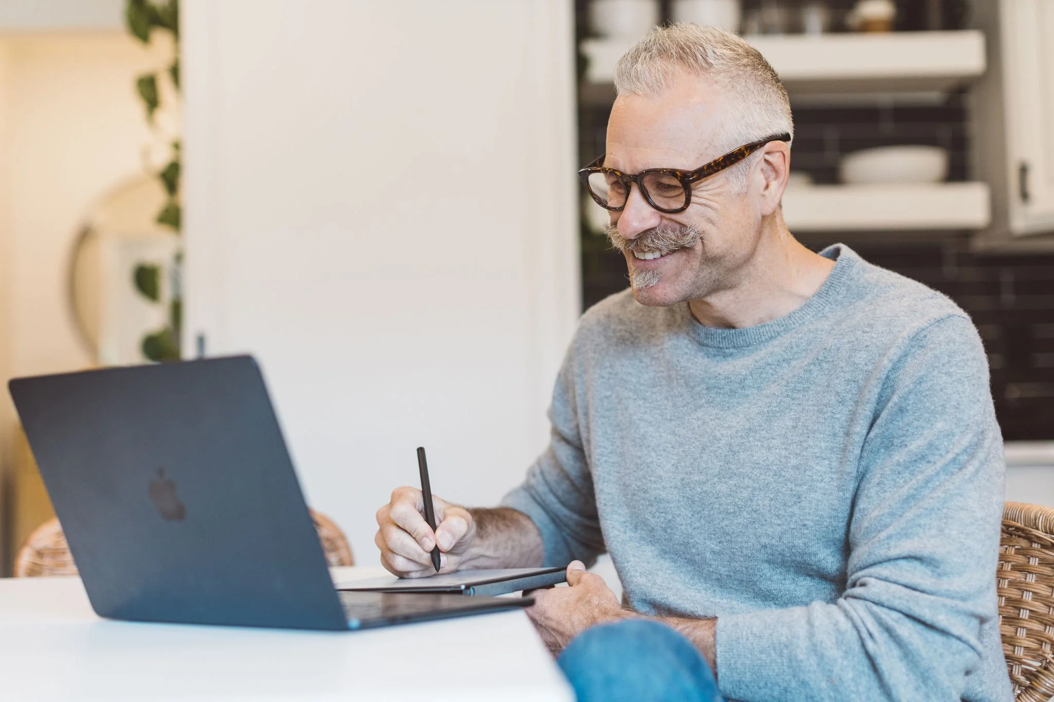 A man with glasses and a mustache, wearing a gray sweater, working on a laptop and using a stylus on a tablet.