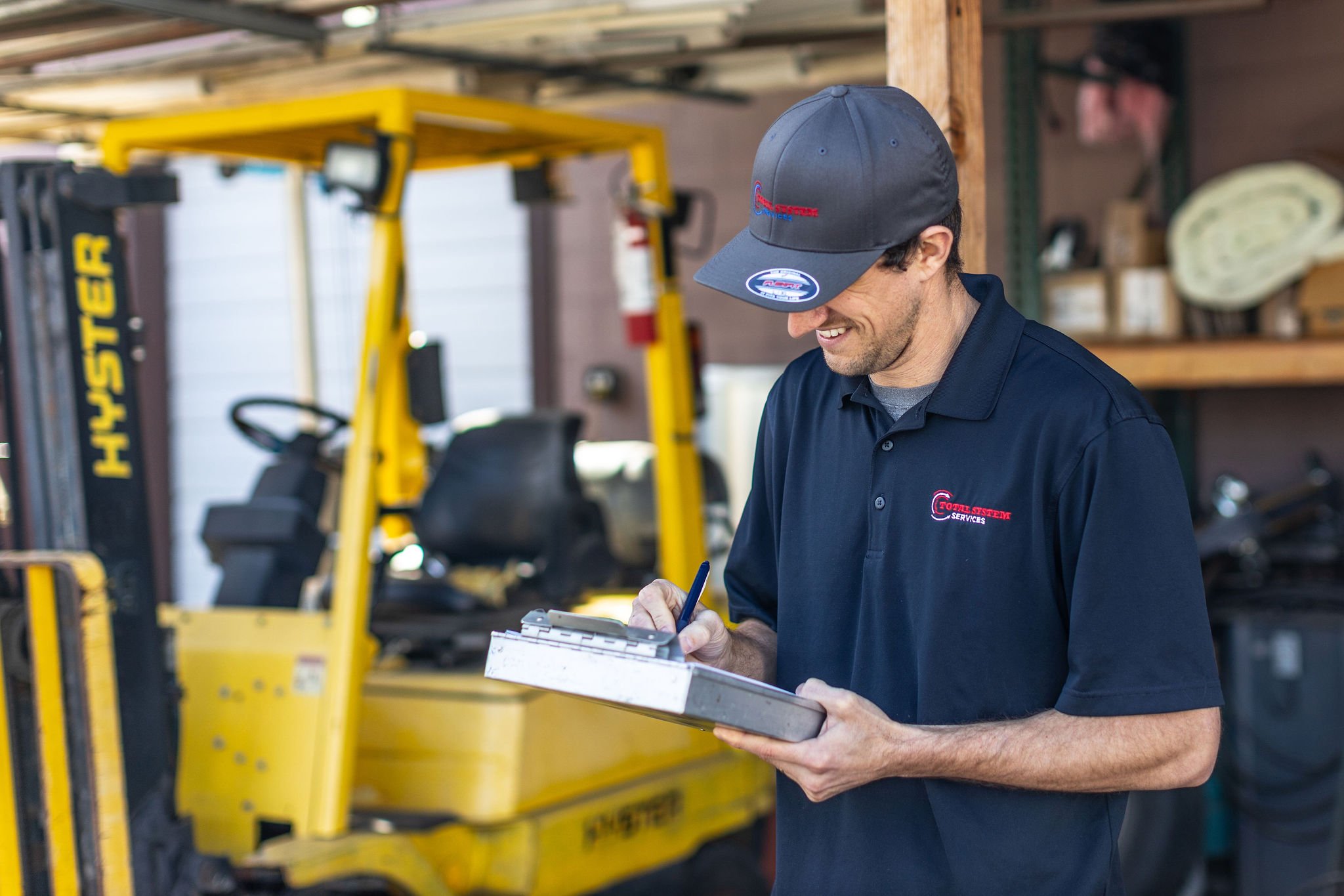 A man wearing a gray cap and navy blue polo shirt is smiling and writing on a clipboard inside a workshop with a yellow forklift and shelves in the background.