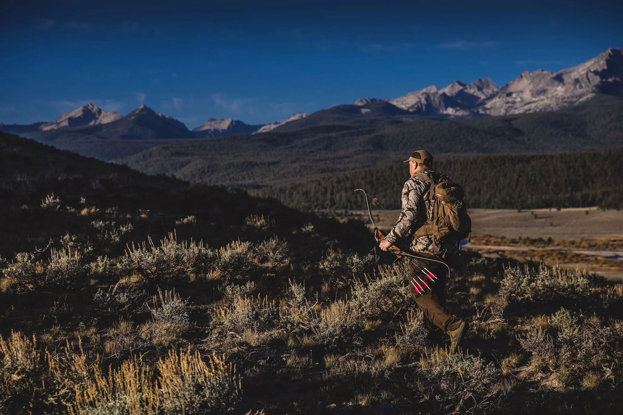 A man in camouflage clothing hiking in a mountainous wilderness area with a bow and arrows, surrounded by shrubs with mountains in the background.