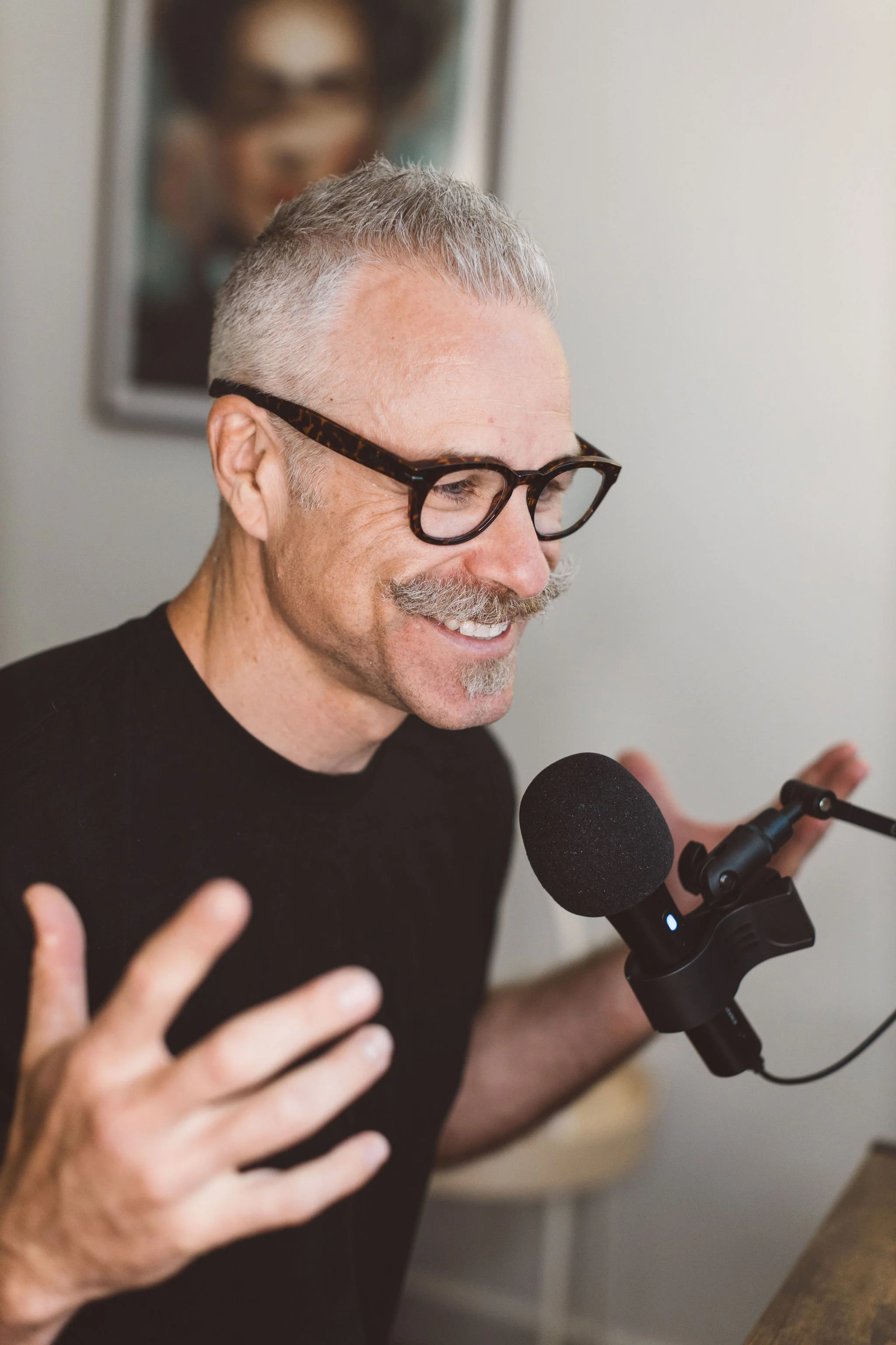 A smiling man with gray hair and a mustache, wearing glasses, speaking into a microphone.