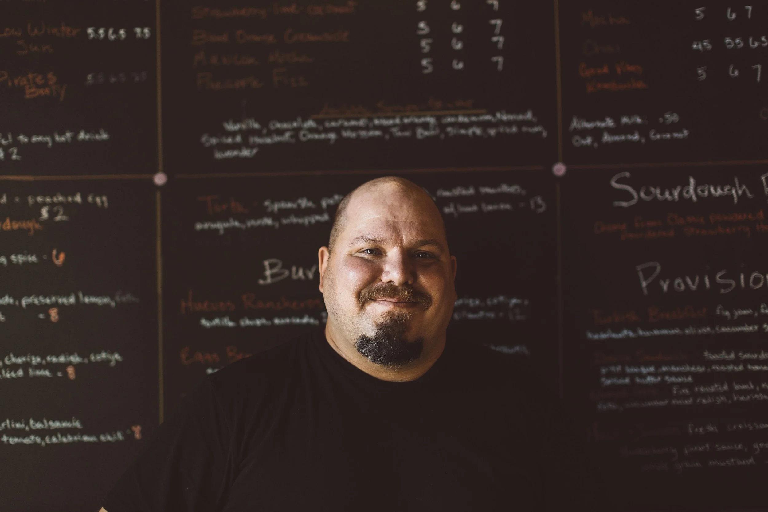 An entrepreneur with a beard and mustache smiling in front of a blackboard menu during a branding photoshoot in Boise, Idaho.