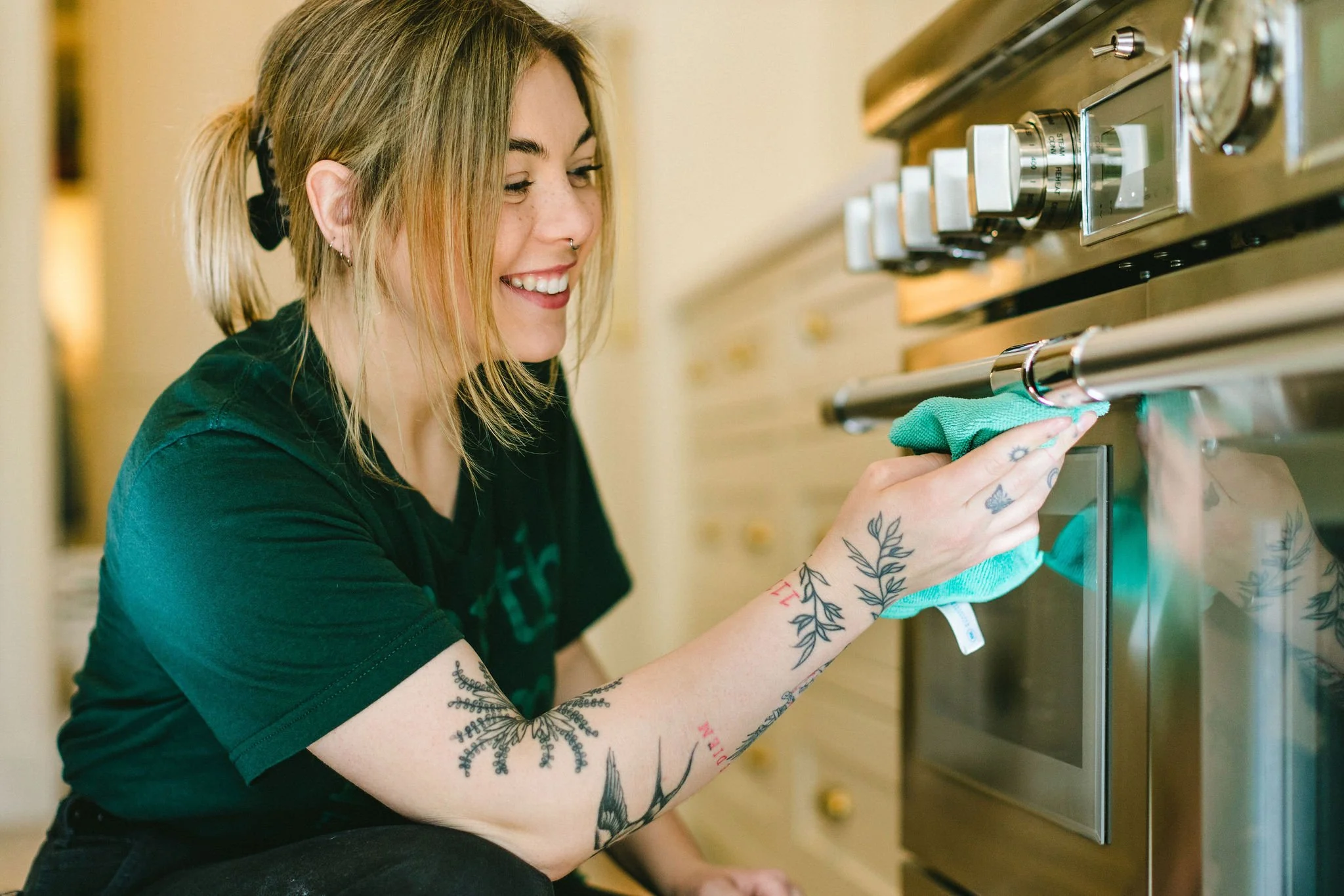 A woman with tattoos on her arms in Boise Idaho, smiling and cleaning an oven with a green cloth during a branding photo session.
